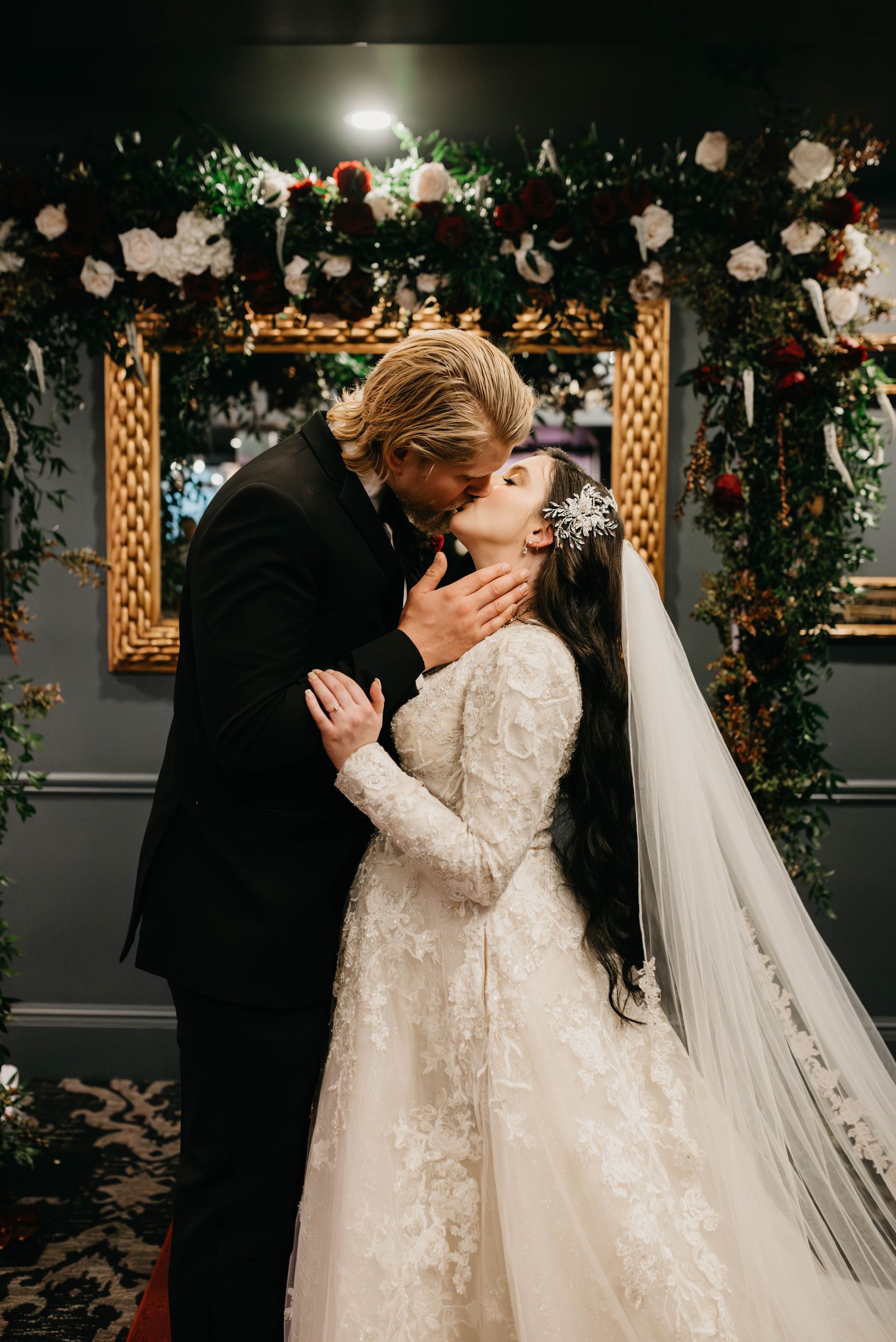 Bride and groom share a kiss beneath floral archway; bride wears white gown, groom in black suit.