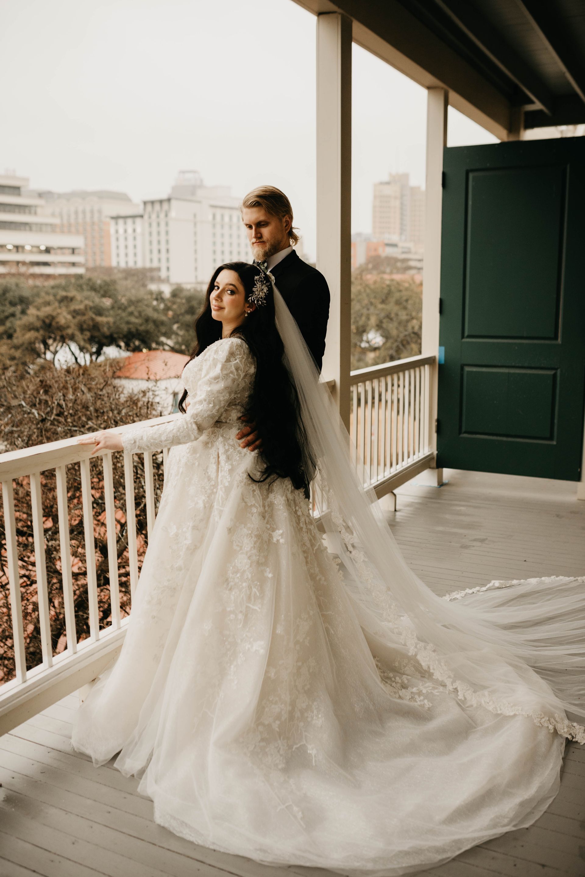 Bride and groom on a balcony overlooking a city. The bride wears a white gown, the groom a black suit.
