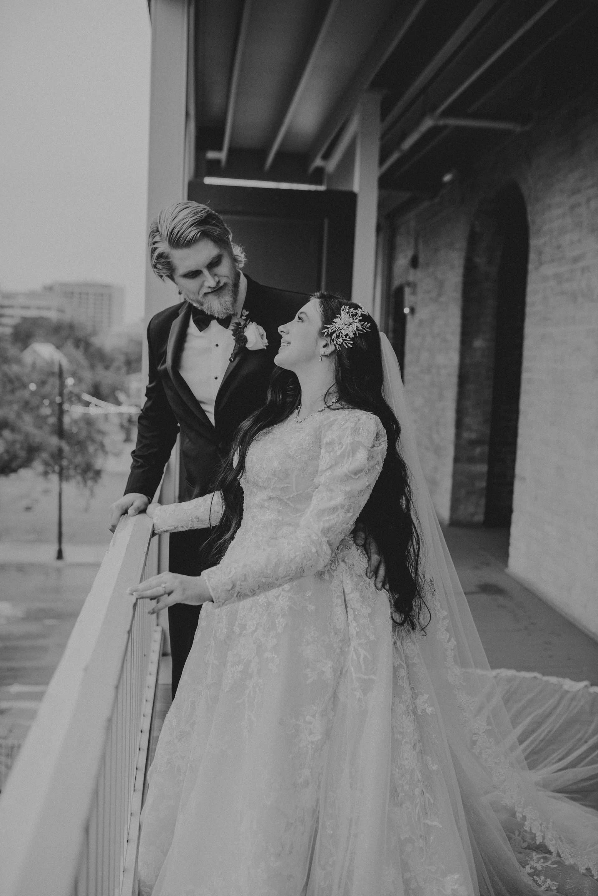 Bride and groom on a balcony, bride in a gown gazes up at the groom who looks down at her.