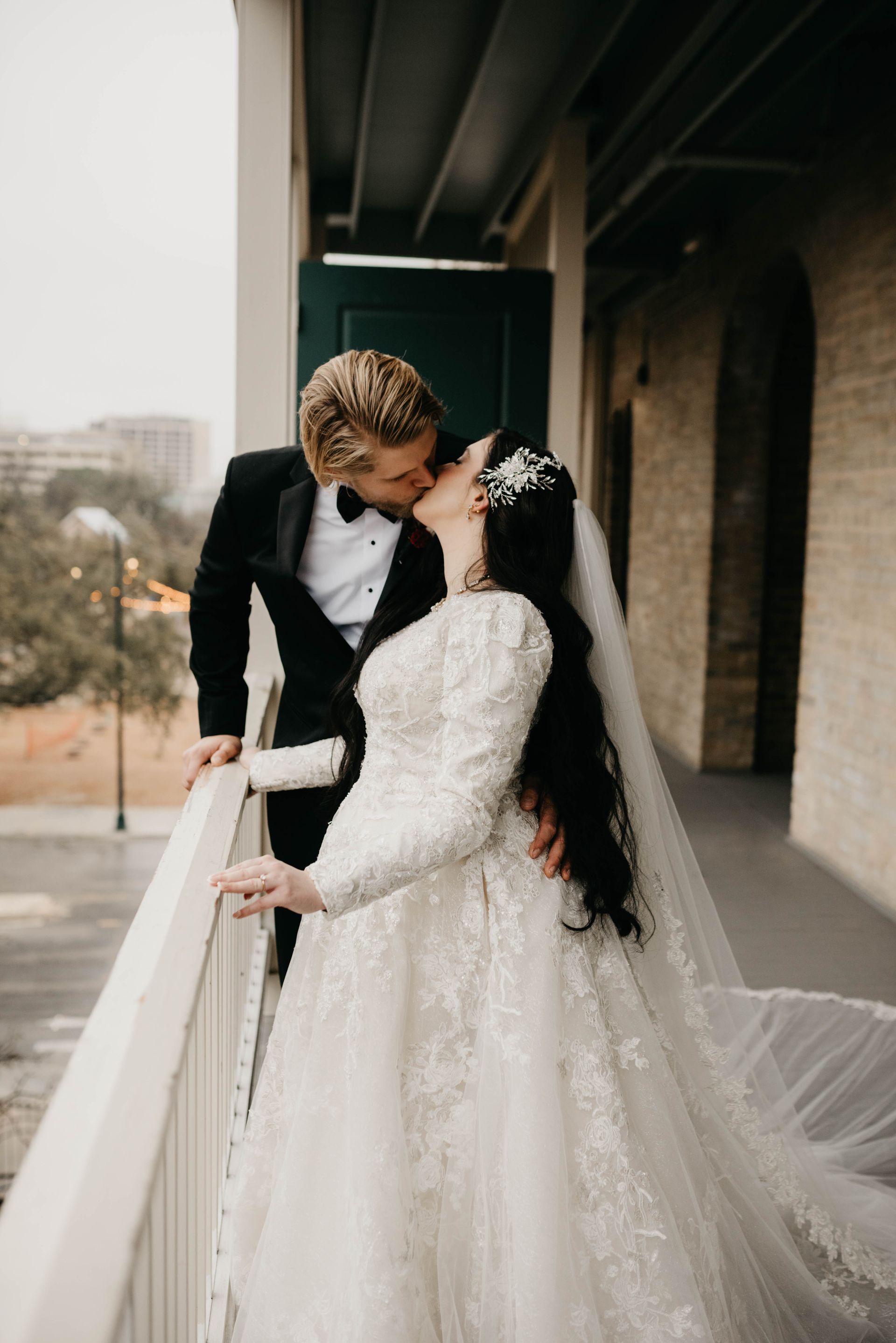 Bride and groom kiss on a balcony. Bride in white gown with veil, groom in tuxedo.