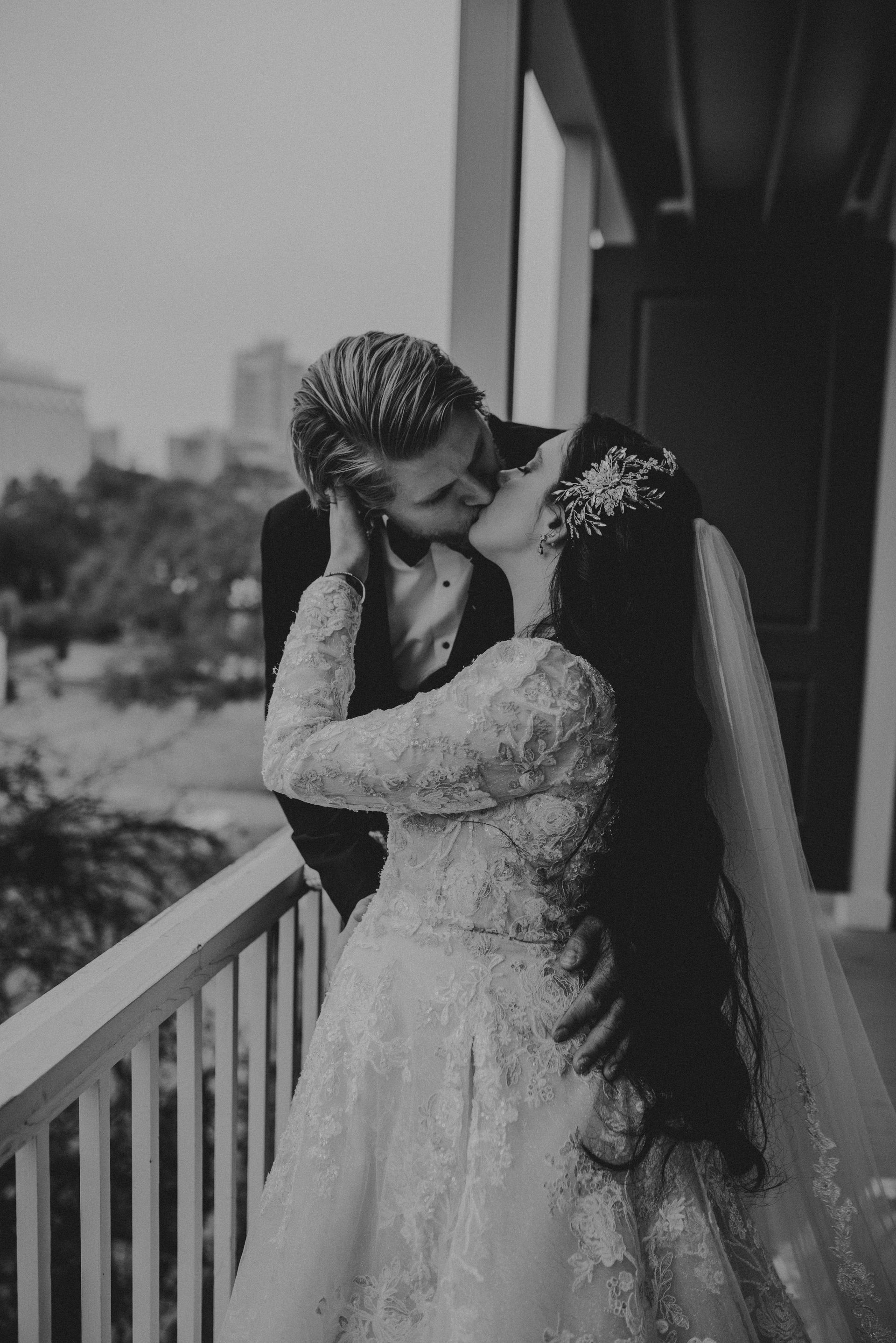 Bride and groom kissing on a balcony. Bride in a long sleeve wedding gown, groom in a suit. Black and white.