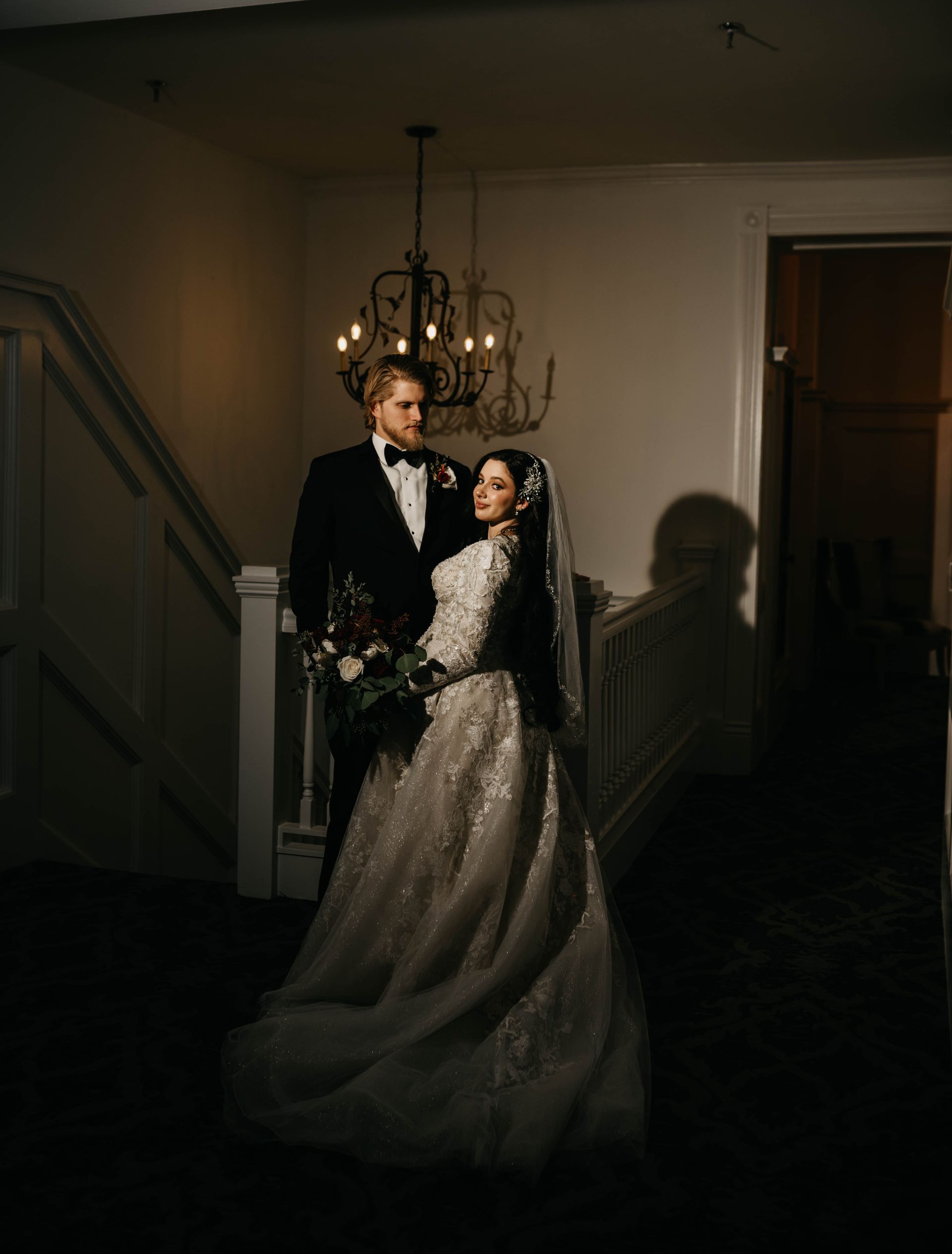 Bride and groom in formal wear, standing by a staircase, illuminated by soft light.
