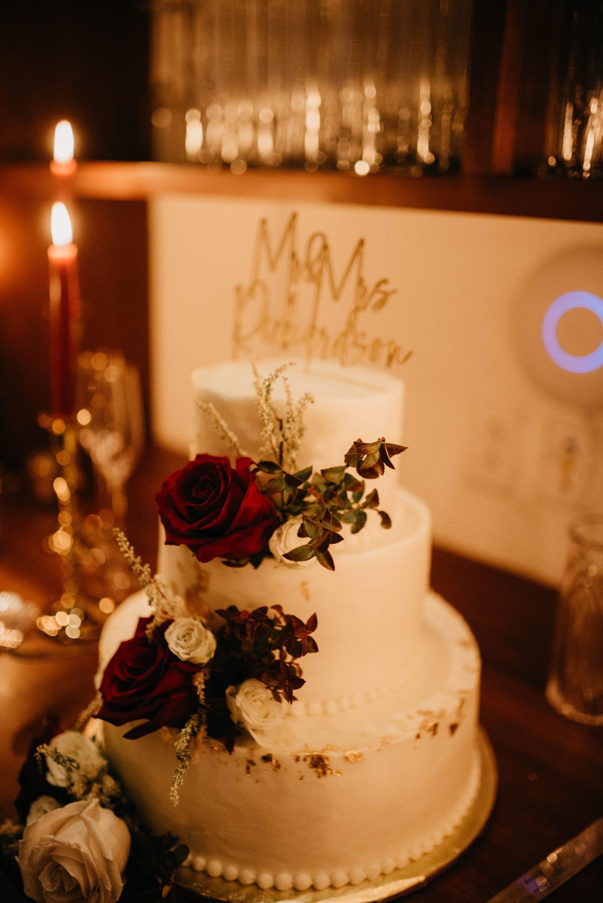 Three-tier white wedding cake decorated with red roses, gold topper, and candles in a dim setting.