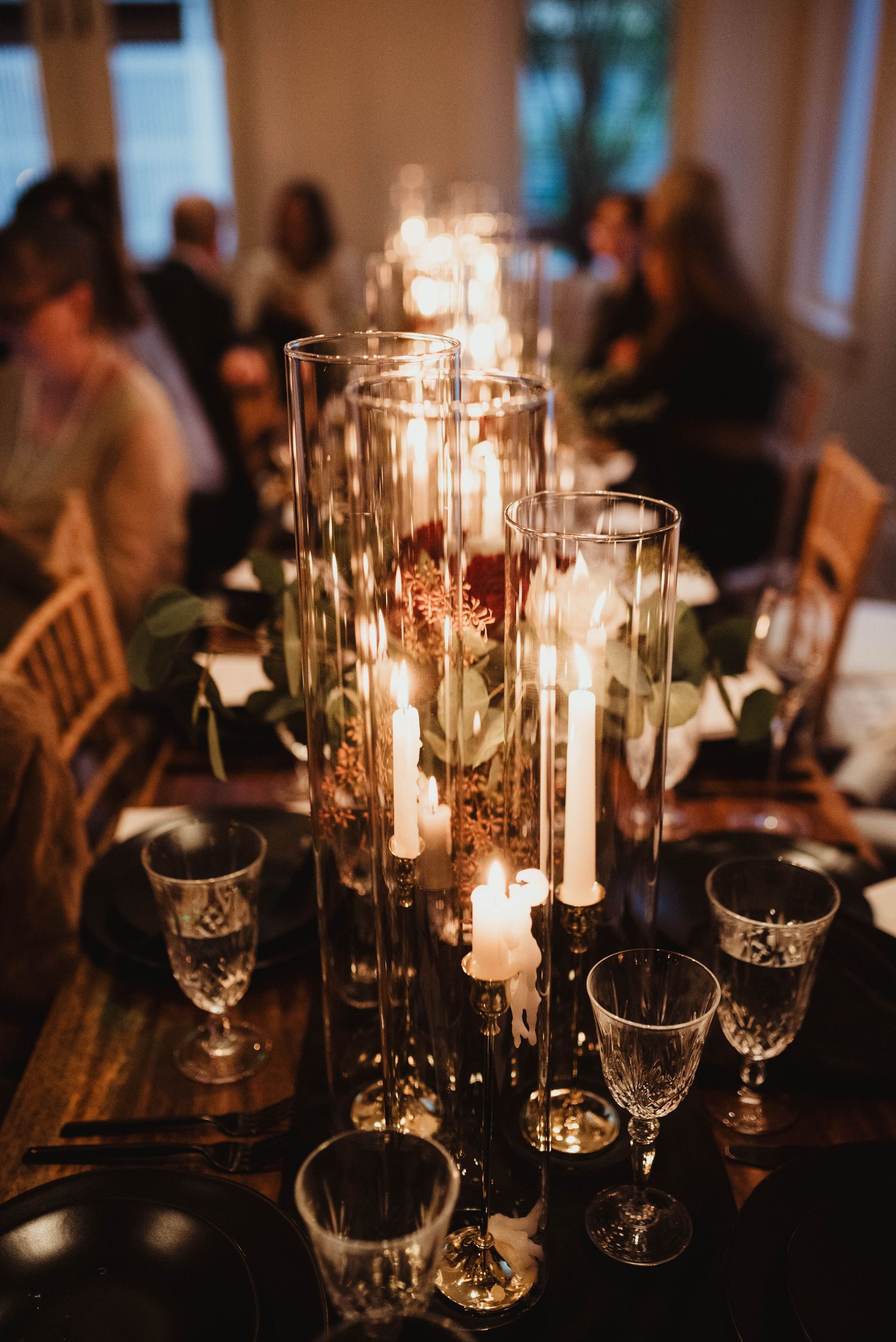 Lit candles in glass cylinders on a decorated table with people in the background.