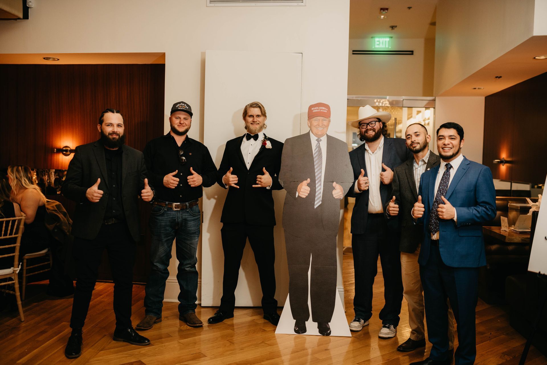 Men pose with a Donald Trump cutout, all giving thumbs up, indoors.