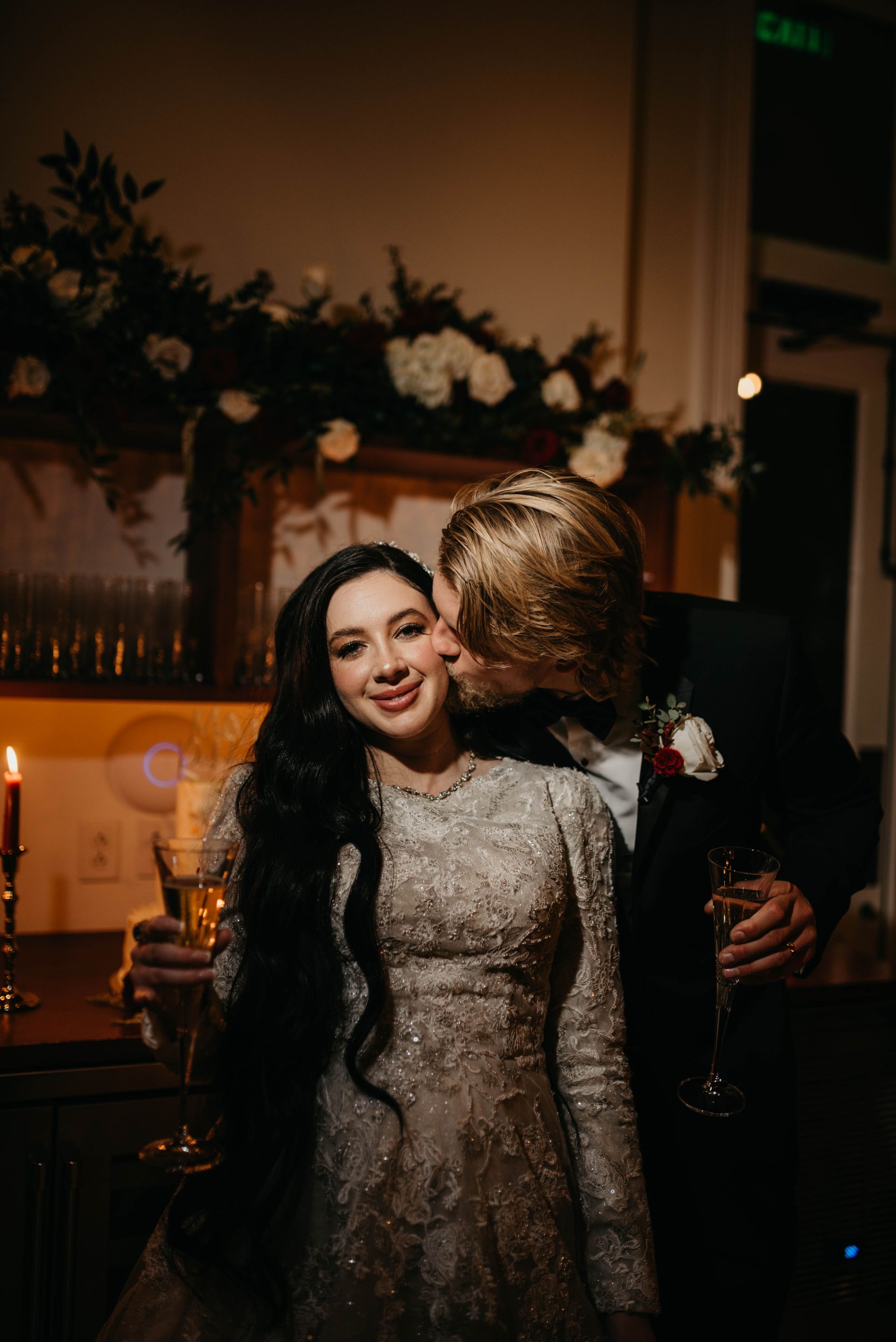 Bride in embellished gown smiles as groom kisses her cheek; floral backdrop.