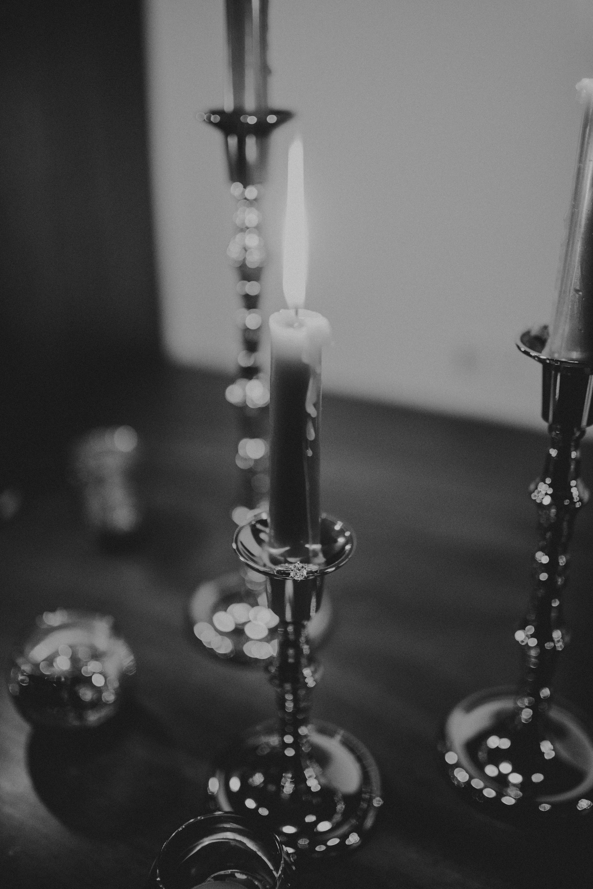 Black and white photo of lit candle in ornate silver candlestick on a dark wooden table.