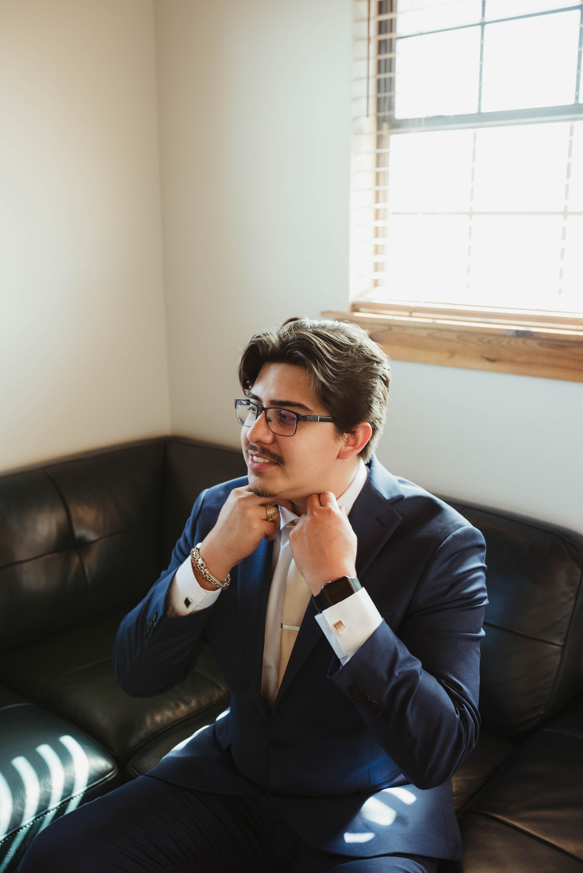 Man in blue suit adjusting bowtie, seated on a black leather couch near a window.