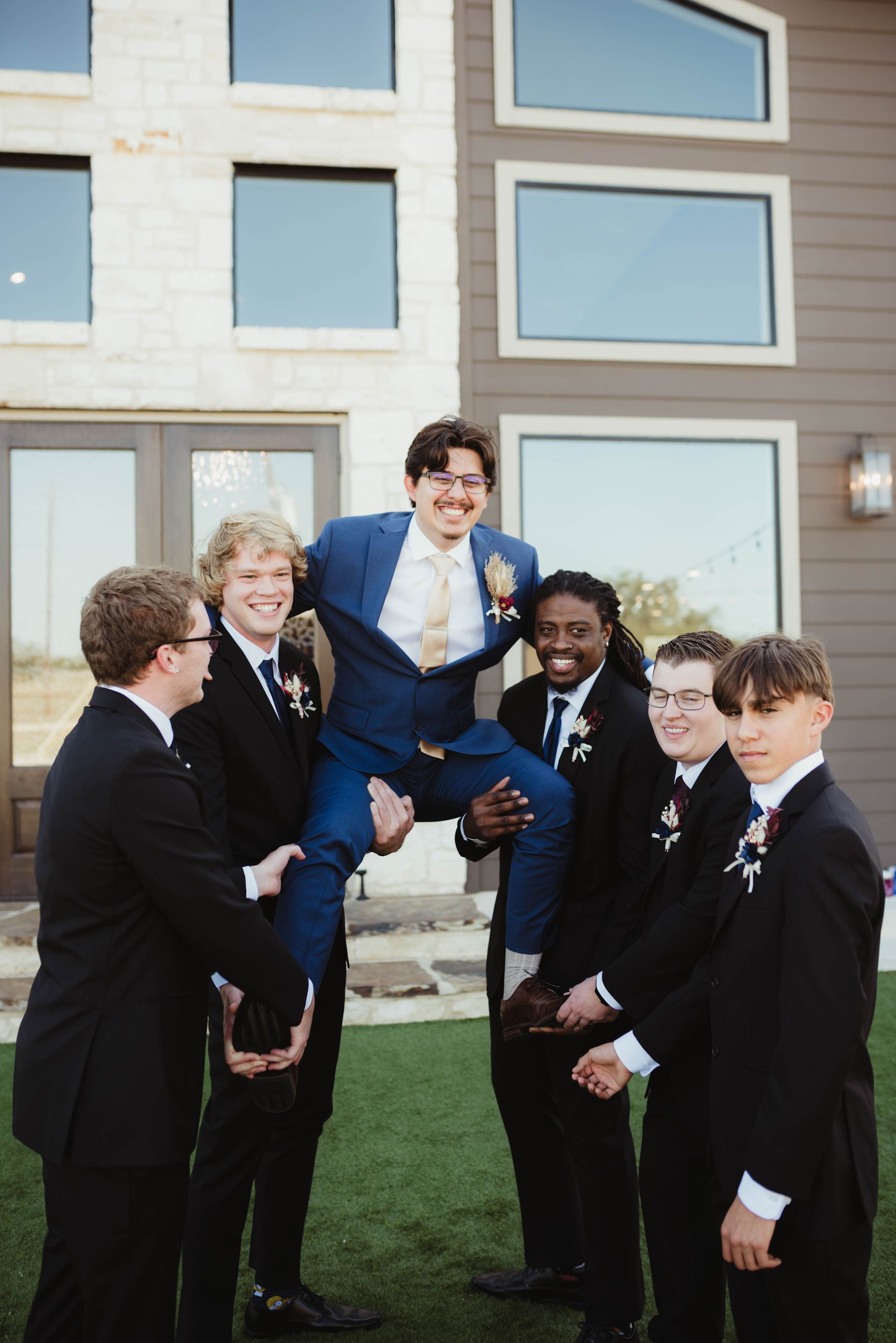 Group of men lifting up another man in a blue suit, outdoors.  Everyone is smiling in front of a house.