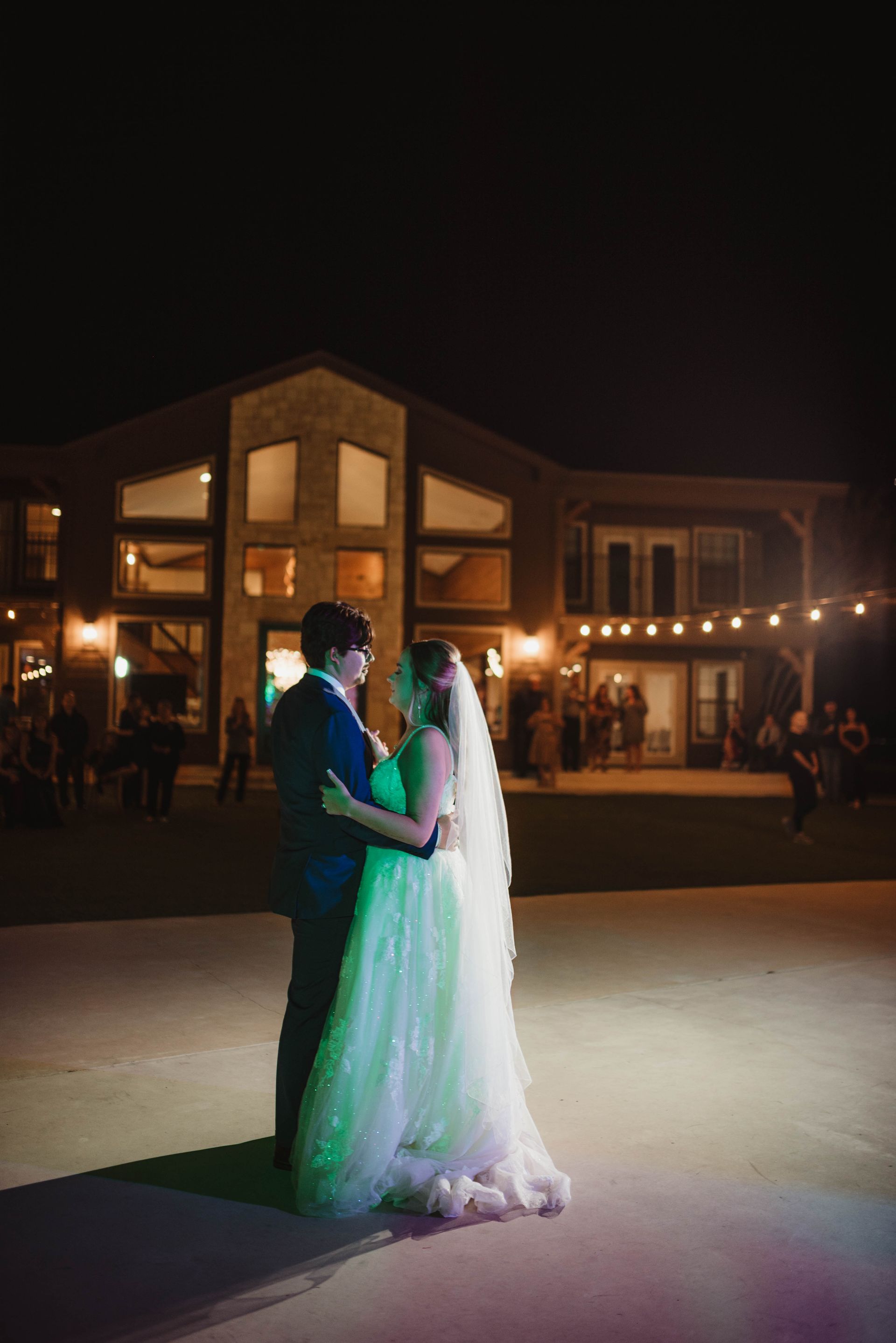 Bride and groom dance on a lit dance floor at night, with a large building in the background.