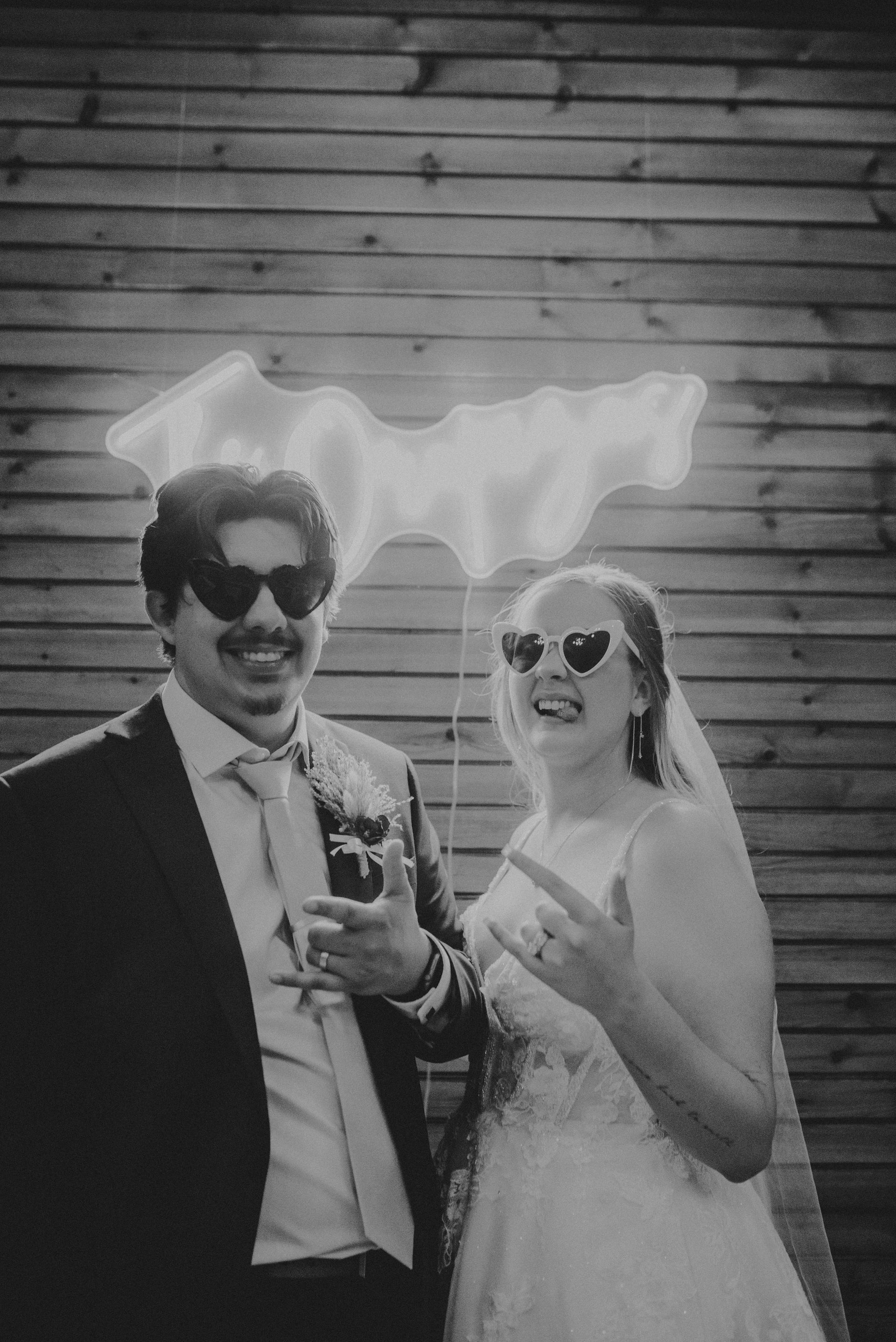 Bride and groom wearing heart-shaped sunglasses, posing for a photo under a neon sign, grinning and making hand gestures.