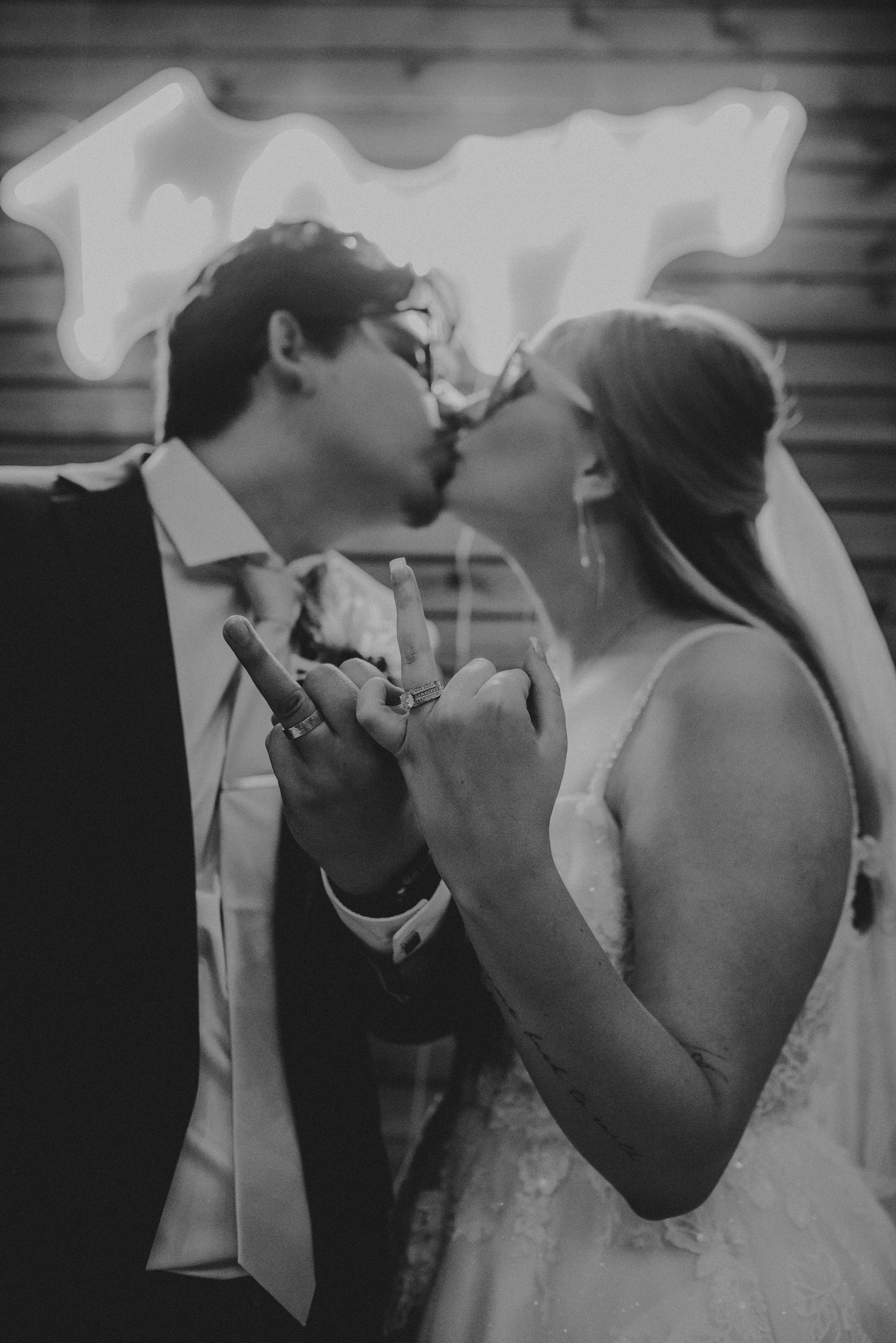 Couple kissing, holding hands, at wedding. Neon sign in background. Black and white.