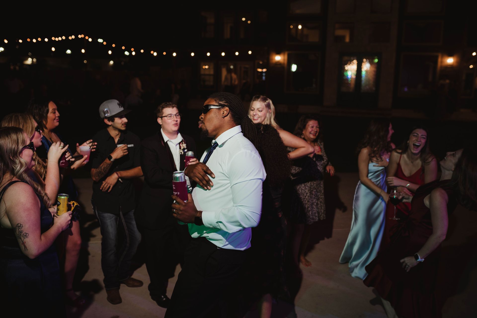 People dancing outdoors at night, holding drinks. String lights illuminate the dark setting.