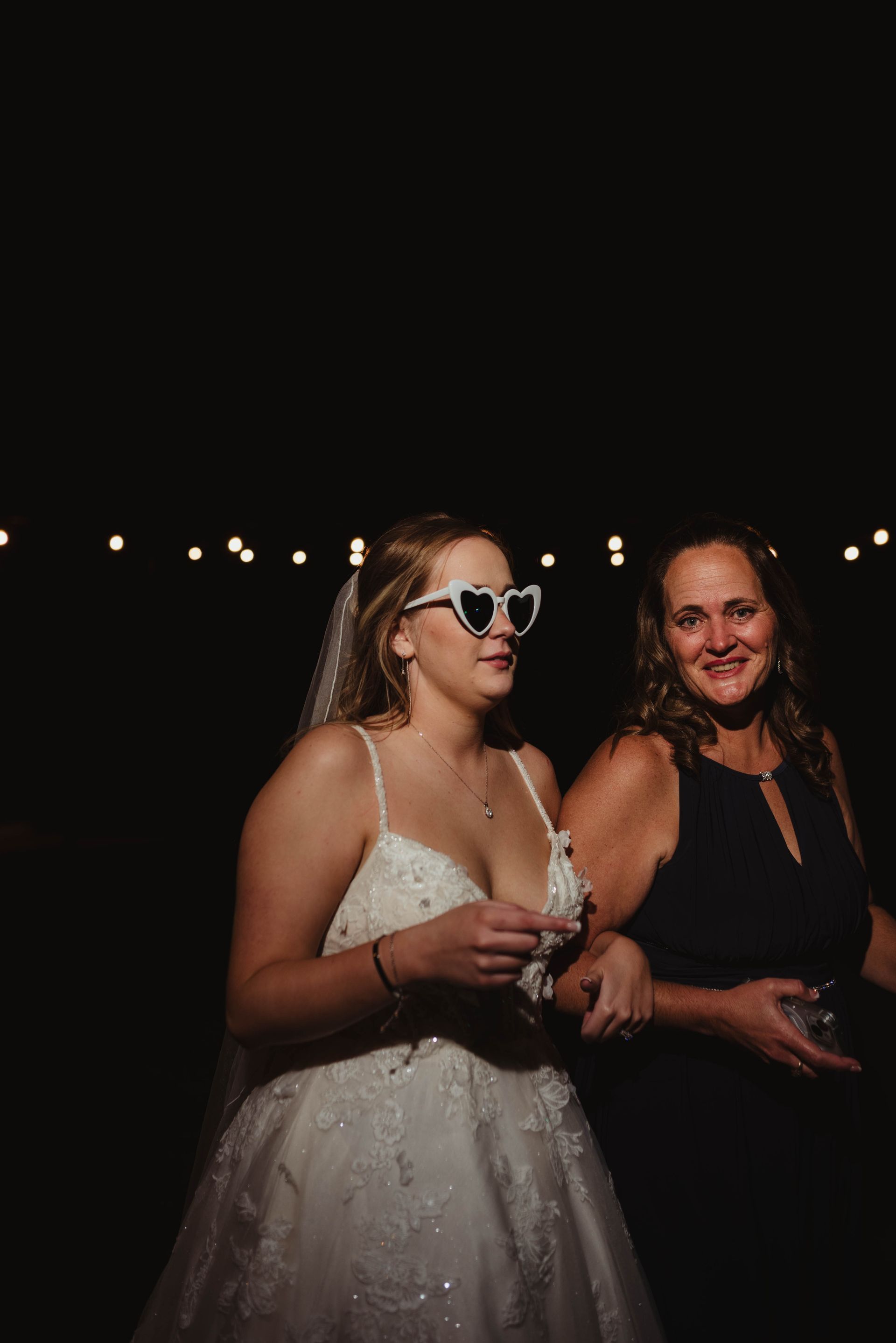 Bride in heart-shaped glasses smiles, arm in arm with a woman in a dark dress, under string lights.