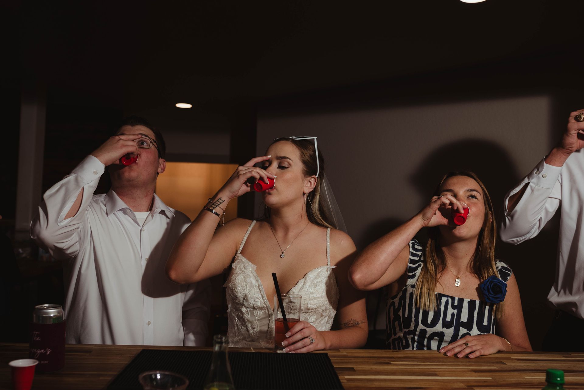 People taking shots at a bar; a bride in a wedding dress is in the center with a veil on her head.