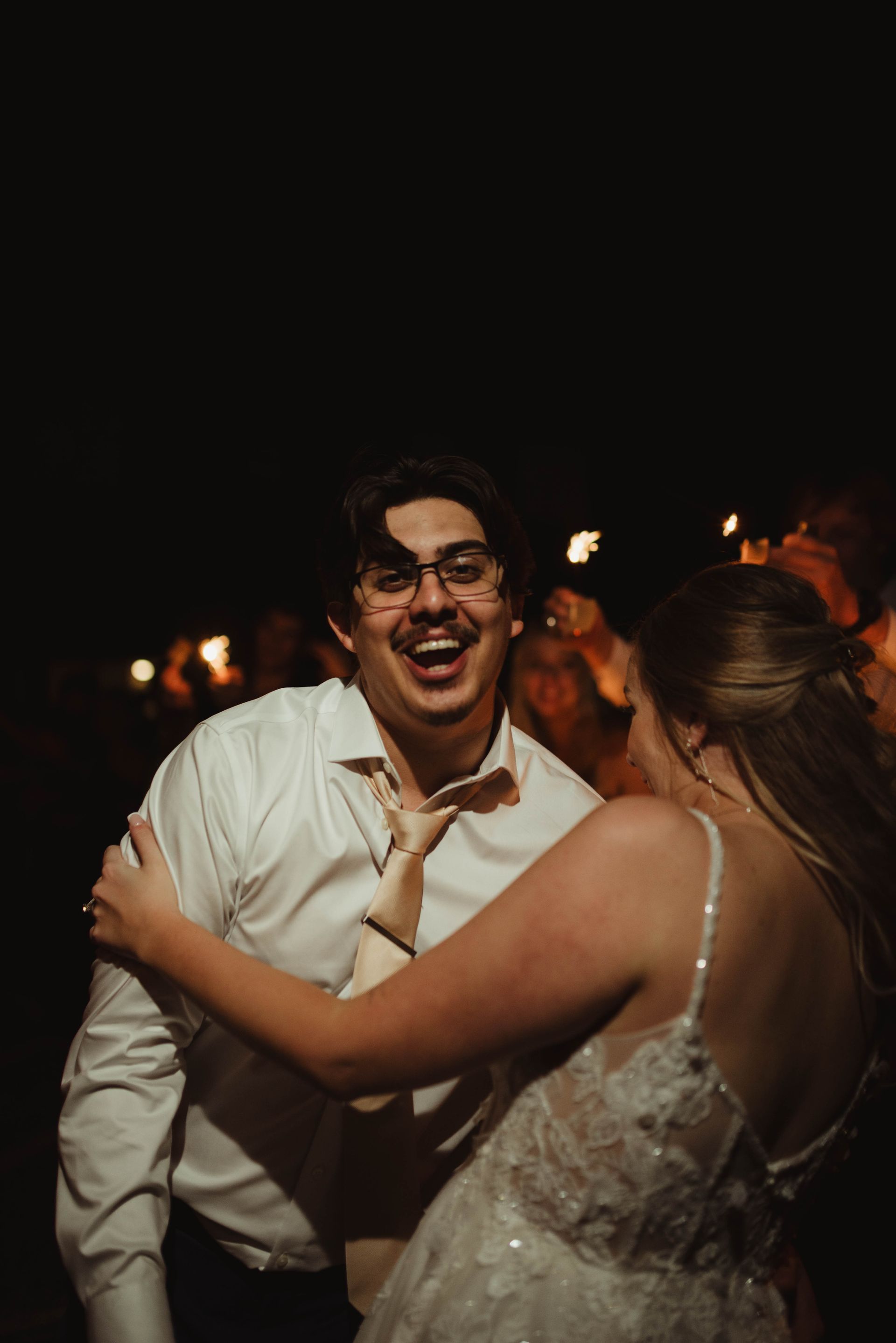 Man with glasses laughs, dancing with a woman in a wedding dress. Night setting.