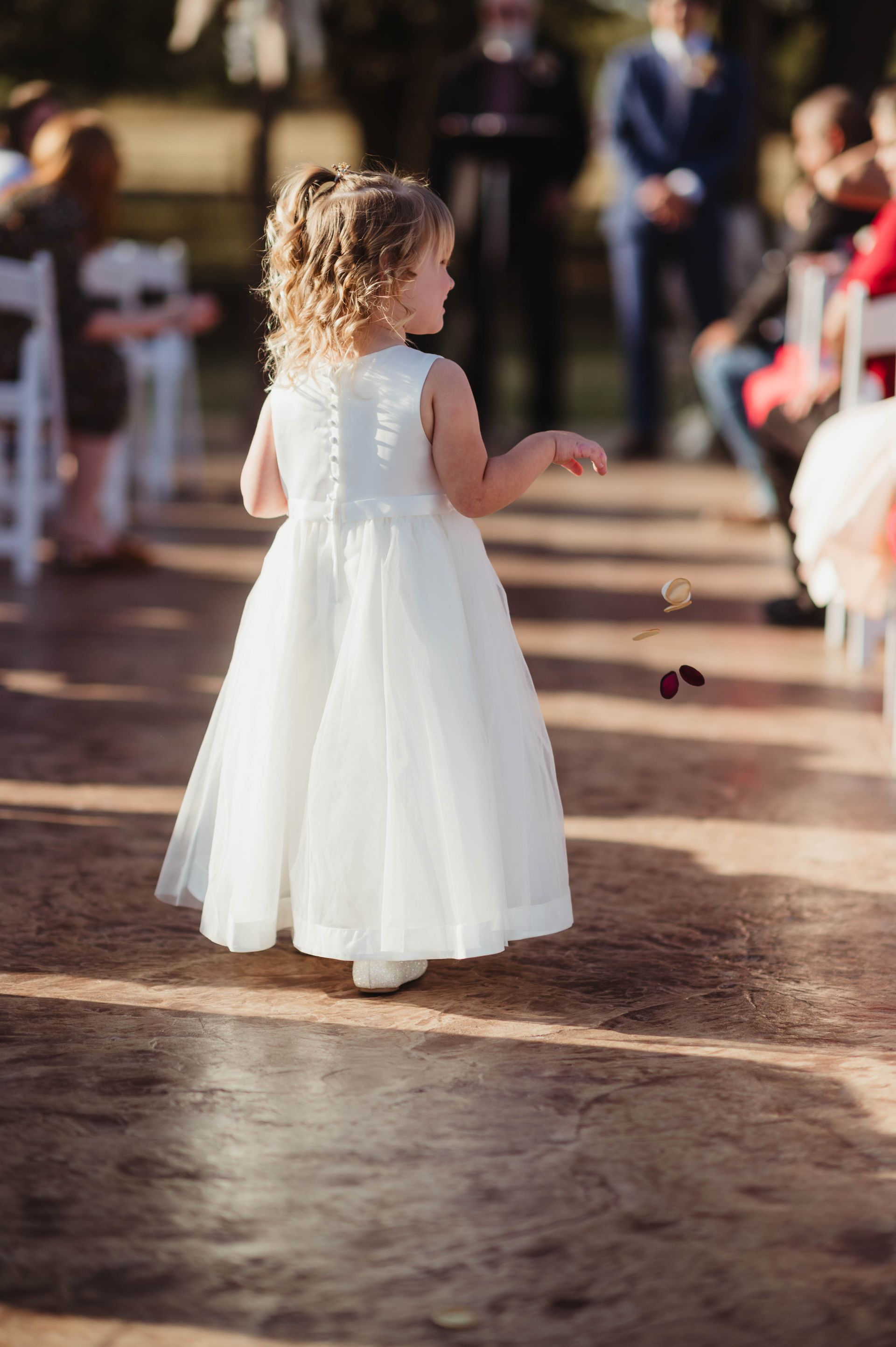 Flower girl in white dress scattering petals at outdoor wedding ceremony.