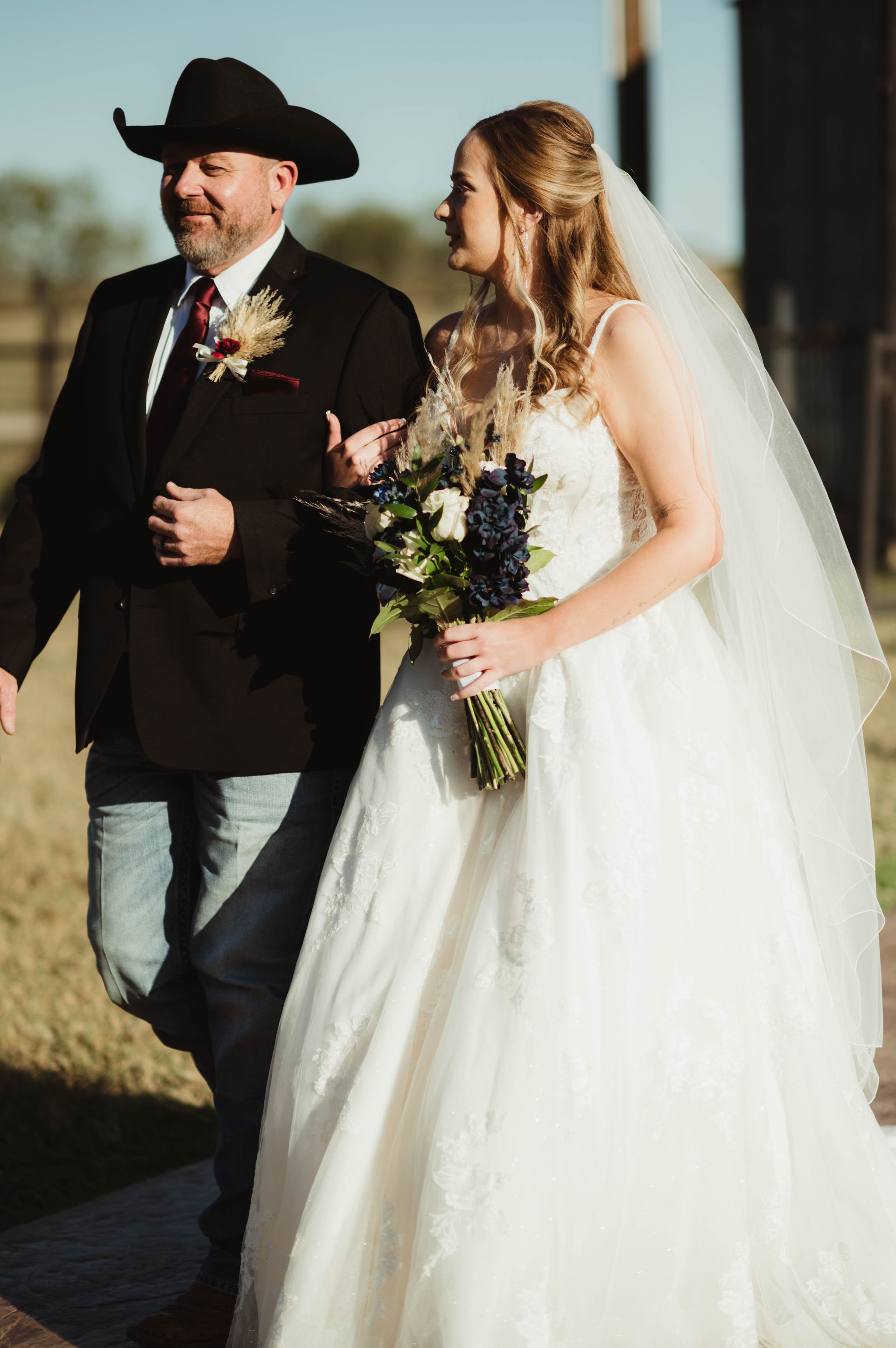 Bride walking with her father; both are outdoors. The bride wears a white dress and veil, her father a black hat and suit.