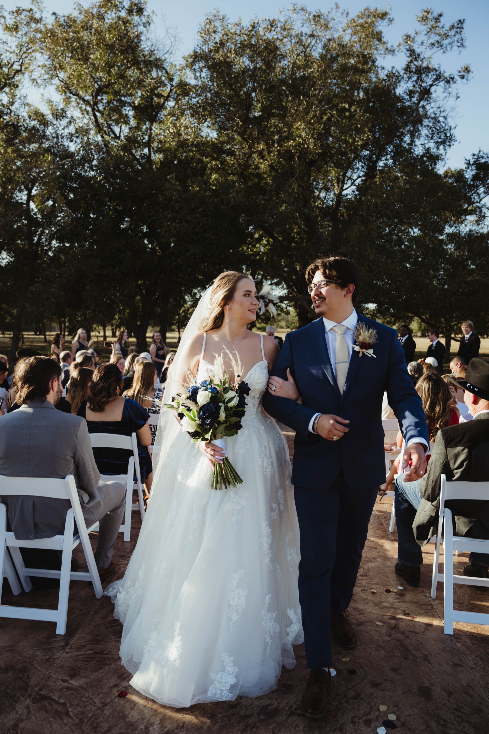 Bride and groom walking, arm-in-arm, after wedding ceremony. Outdoor setting with guests seated.