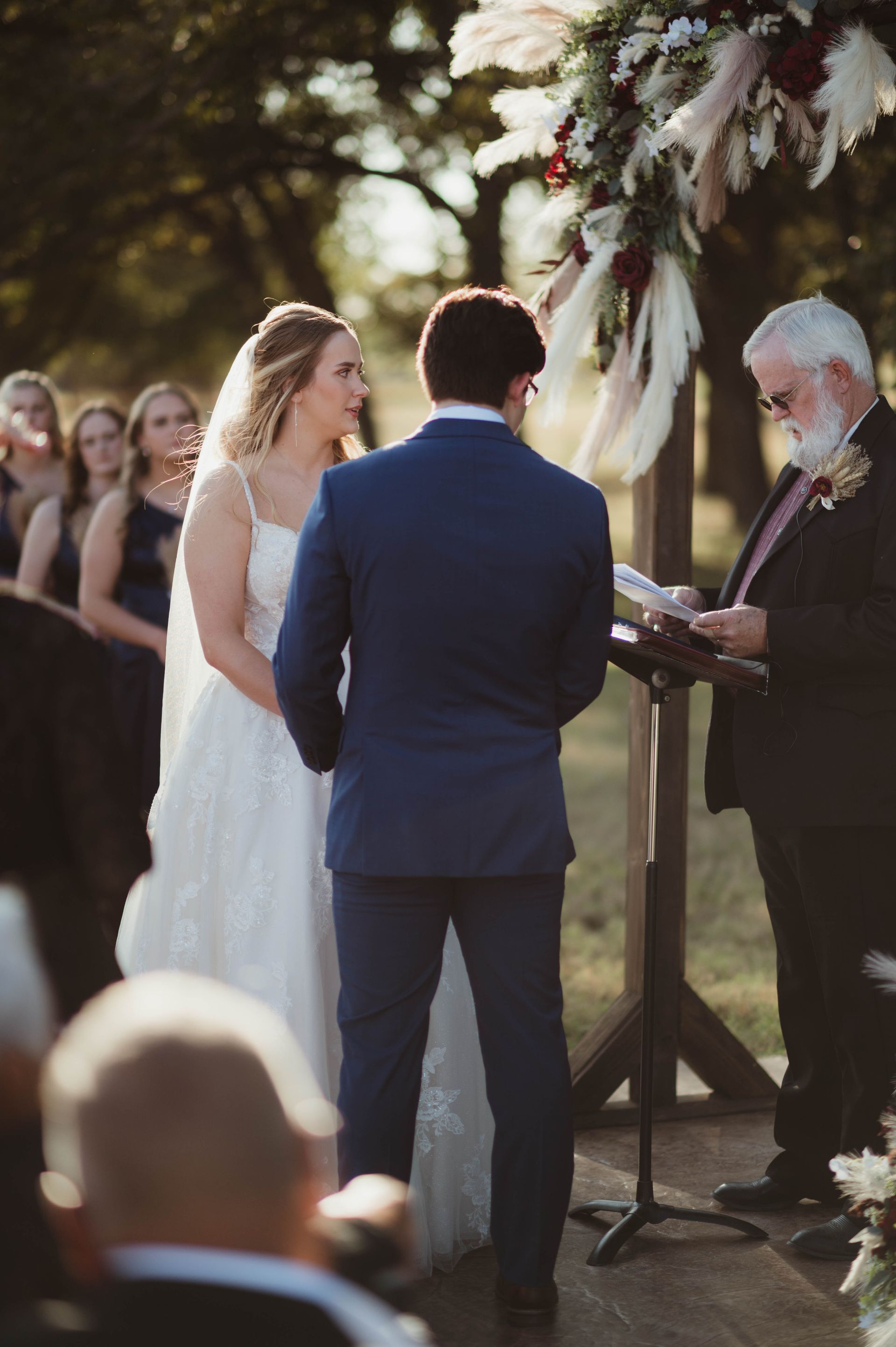 Bride and groom at outdoor wedding ceremony. Wooden arch decorated with flowers. Minister reads from a book.