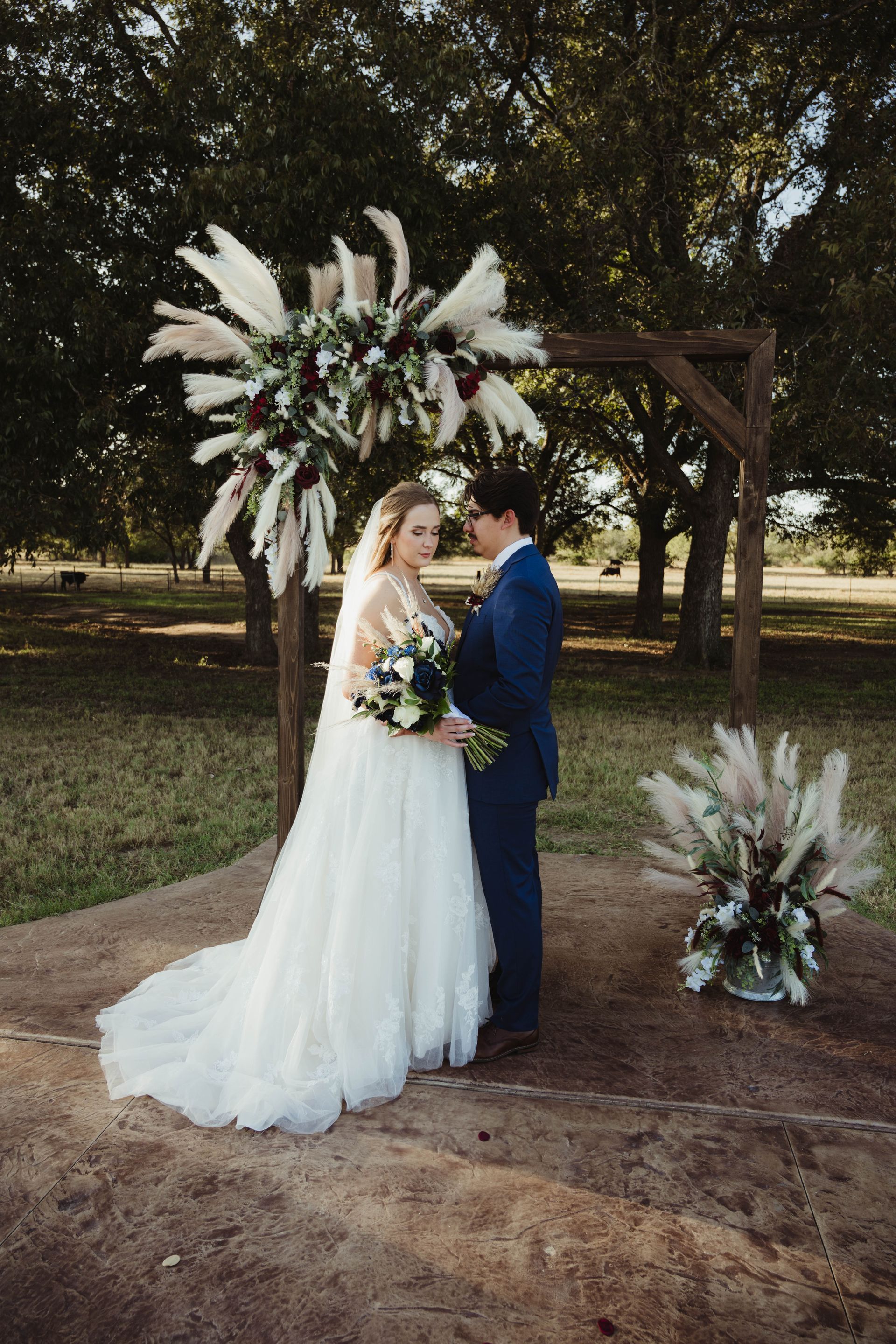 Bride and groom at outdoor wedding ceremony; floral arch and bouquet; trees in background.