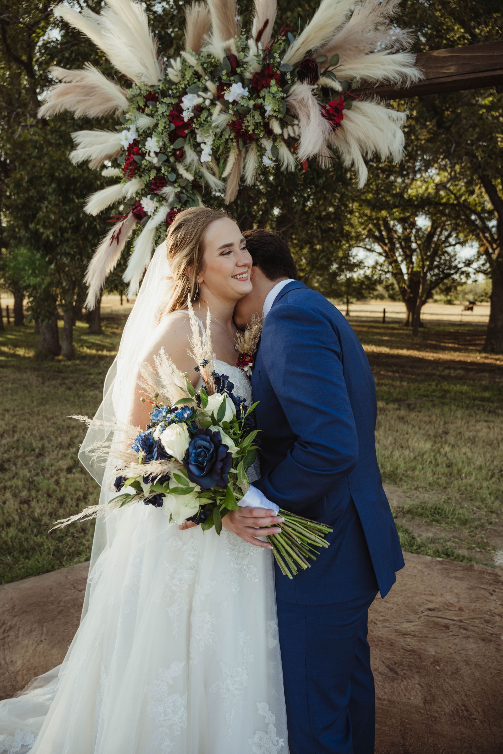 Bride in white dress holds bouquet as groom kisses her cheek under floral arch.