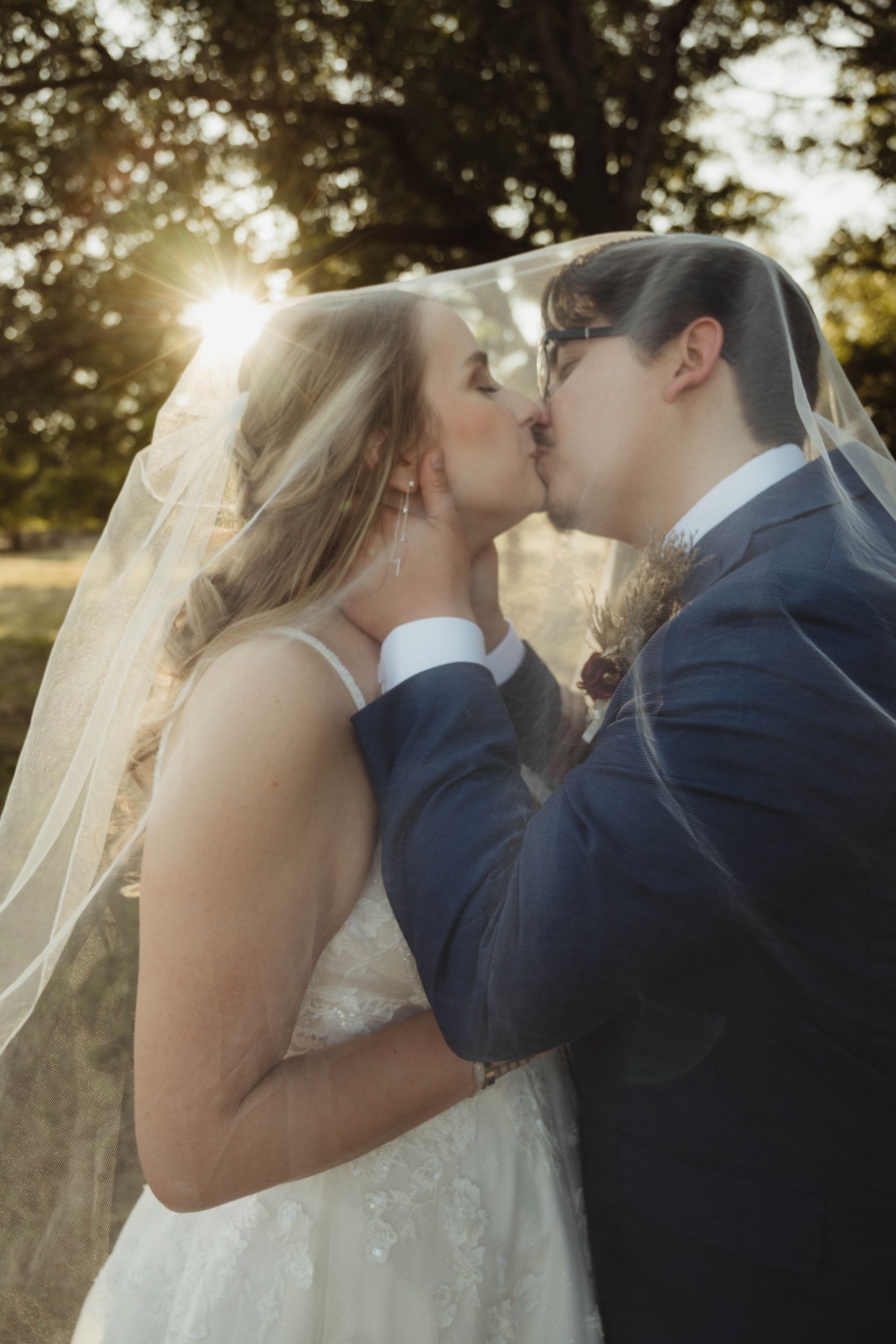 Couple kissing under a veil outdoors, sunlight beaming through trees.