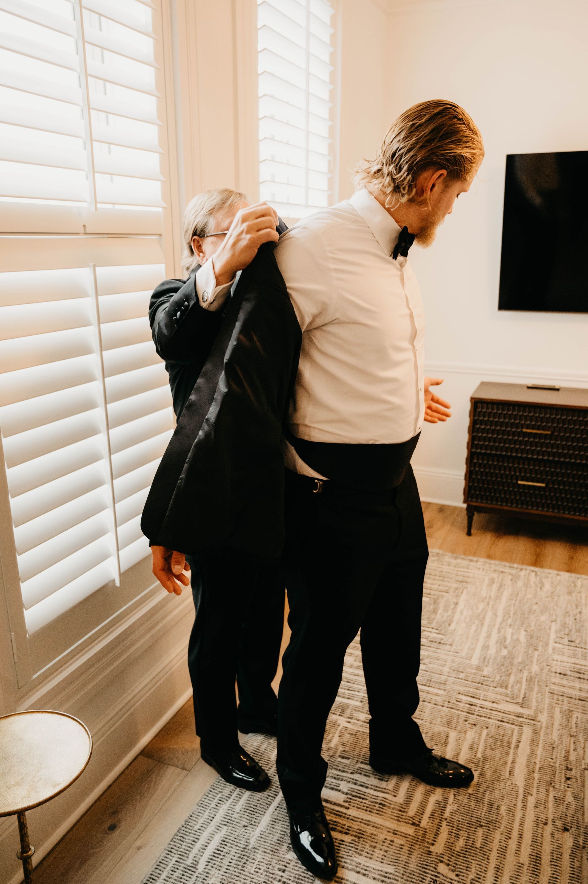 Man helping another man put on a black suit jacket; indoors, near a window with blinds.