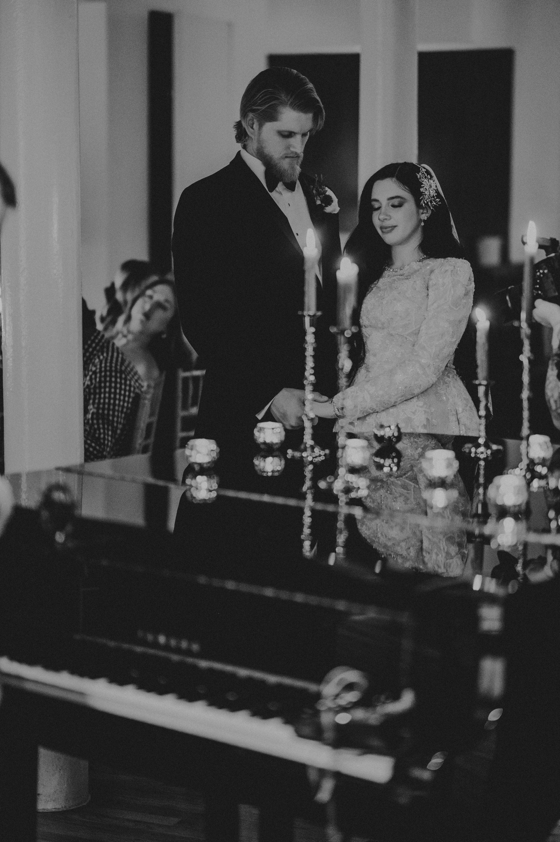 Bride and groom at ceremony, holding hands. Black and white photo, candles and piano present.