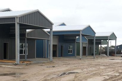Row of unfinished blue metal buildings on a muddy construction site under a gray sky