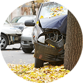 Damaged car crashed into a tree with other wrecked cars in the background on a leaf-covered street.