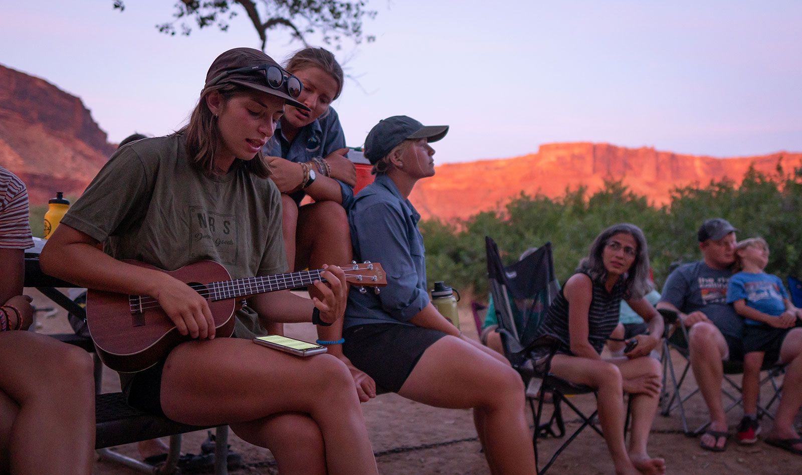A group of people are sitting around a campfire playing ukuleles.