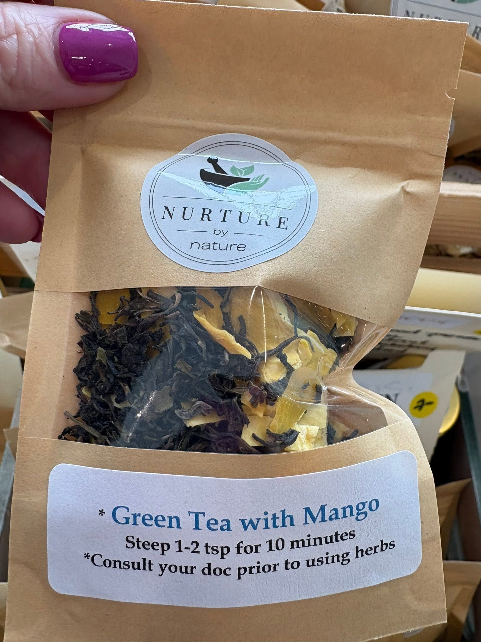 Hand holding a brown pouch of green tea with mango, showing dried tea leaves and mango pieces through the window