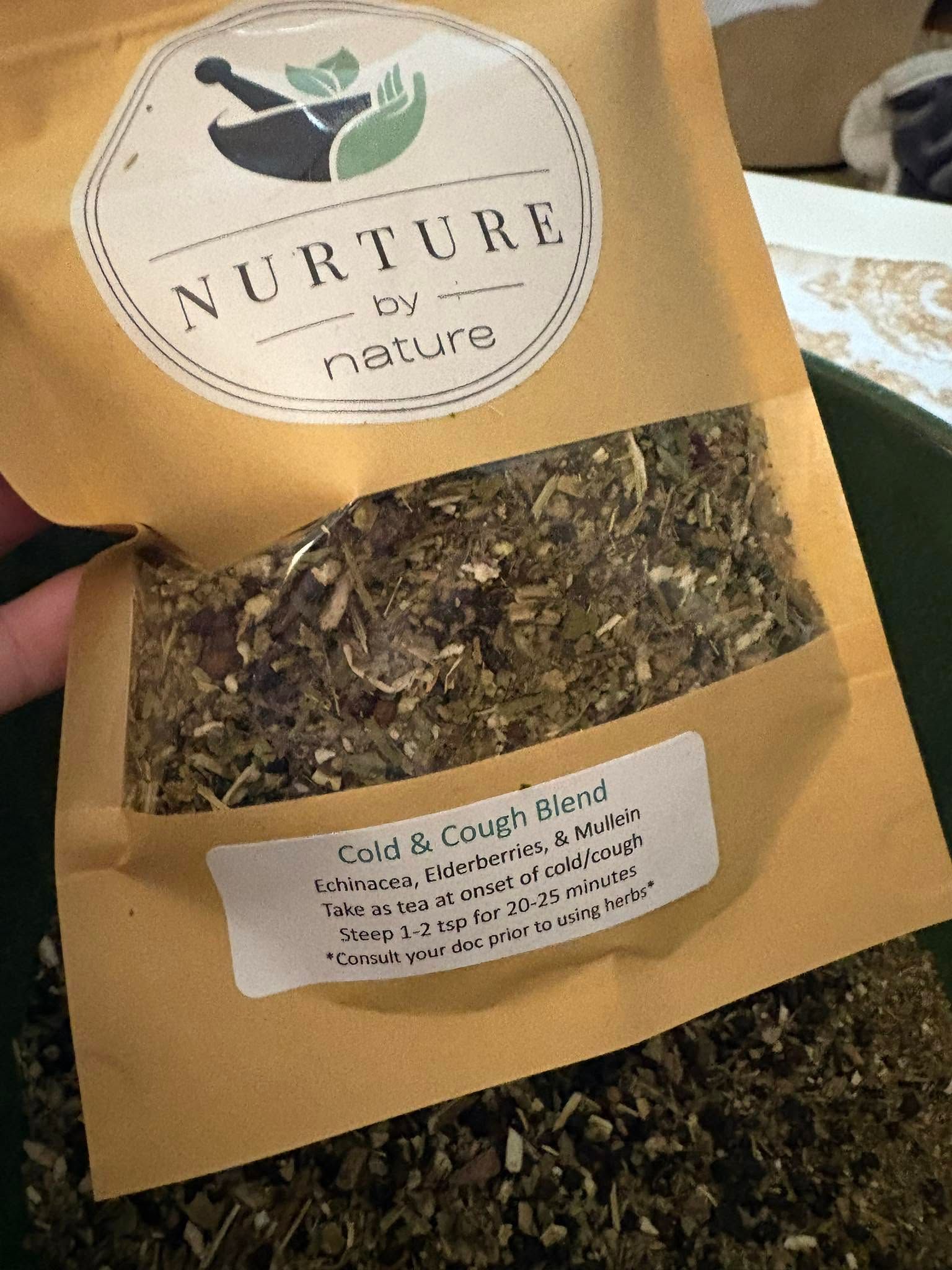 Hand holding a tan pouch of dried tea leaves with a clear window and Nurture Tea label.