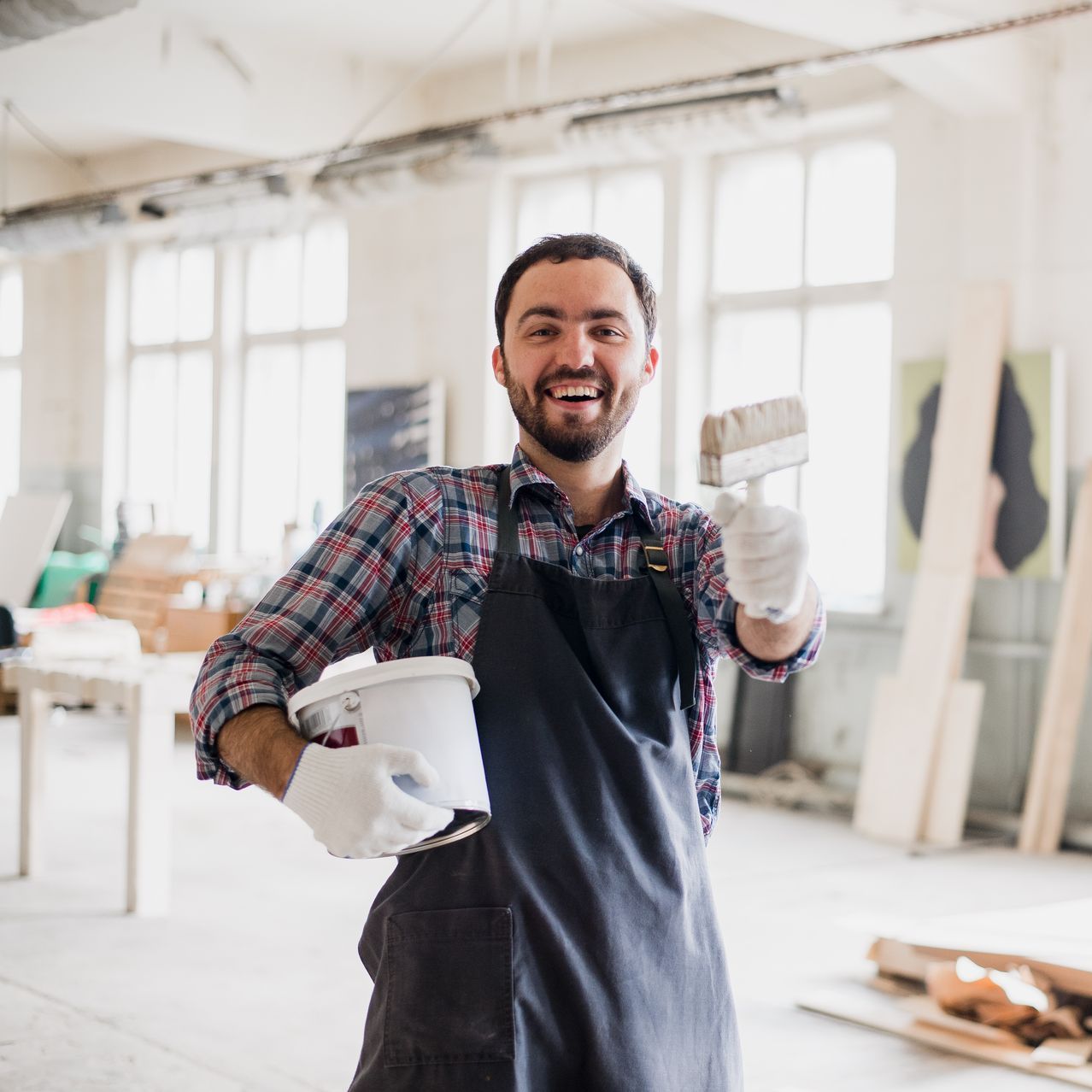 A man in an apron is holding a bucket of paint and a paintbrush.