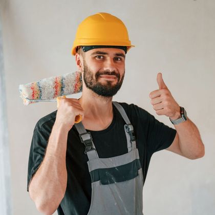A man wearing a hard hat is holding a paint roller and giving a thumbs up.