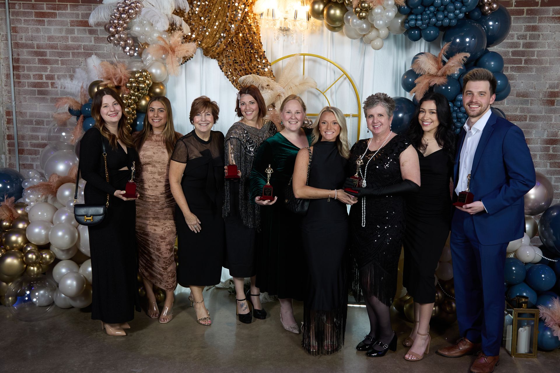 Group of people posing for photo, holding awards at an event. Balloons and decorated backdrop present.