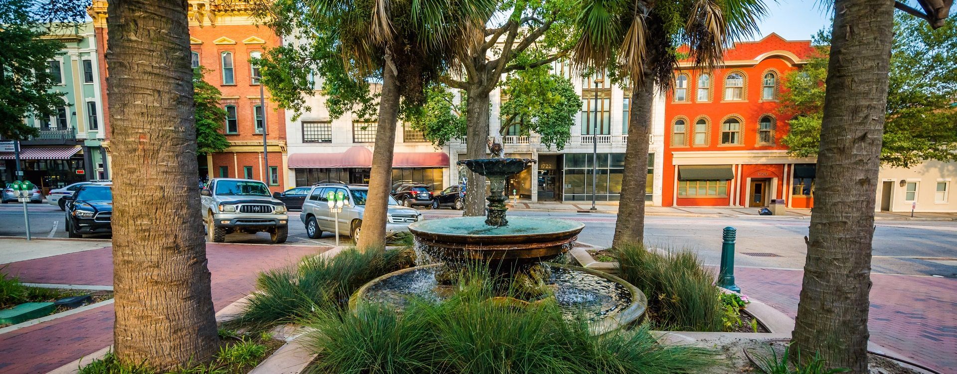 A fountain surrounded by greenery sits in a town square with colorful buildings and palm trees.