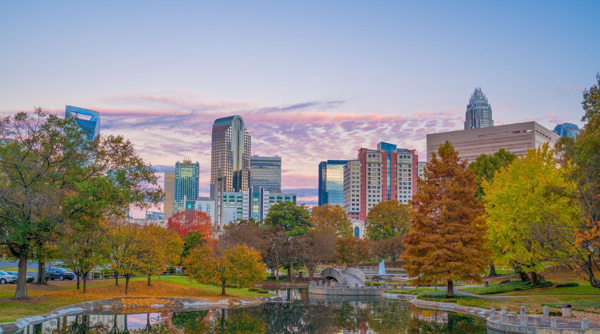 City skyline behind a park with trees and a pond at sunset.