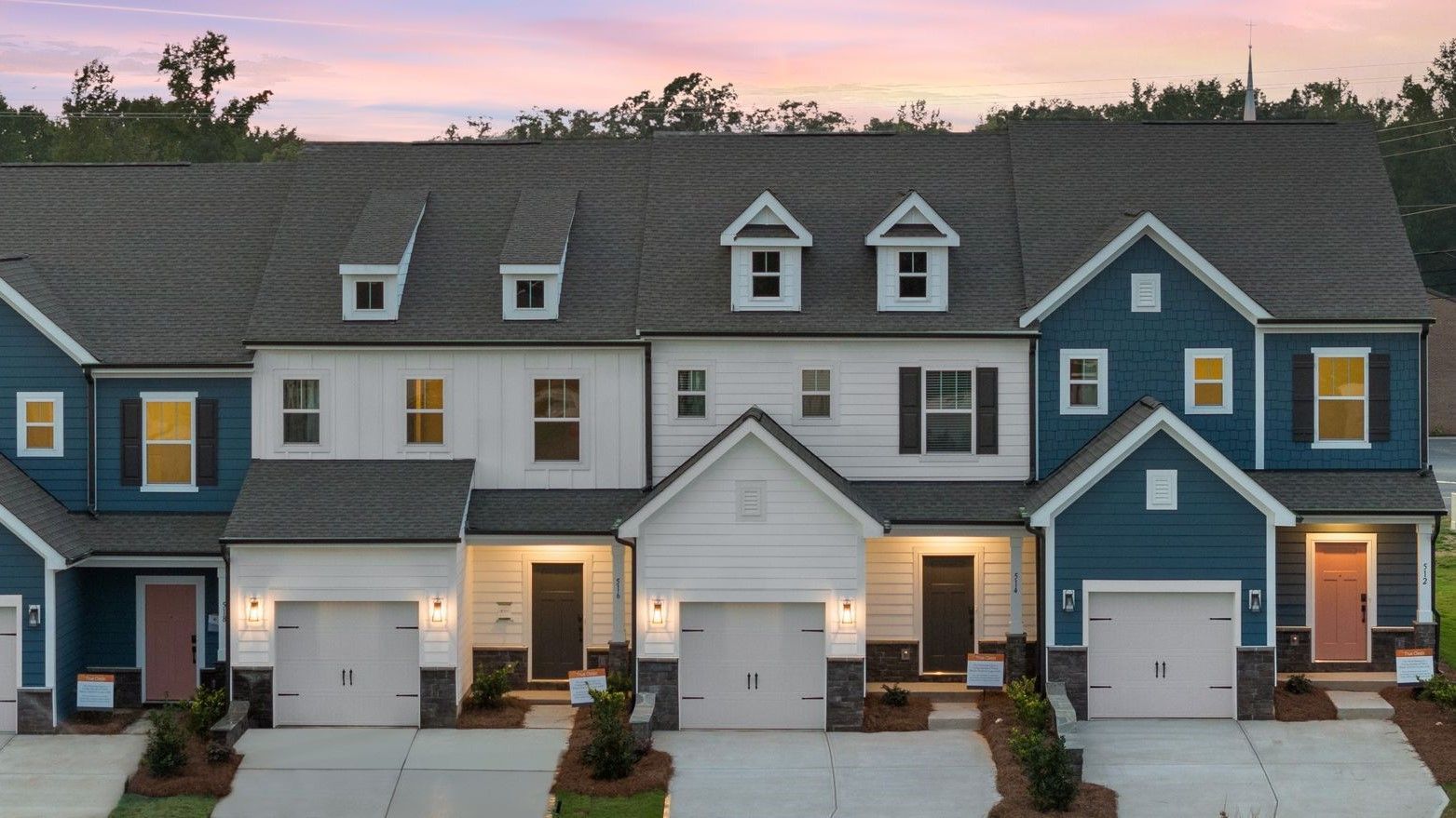 Row of townhouses with dark gray roofs, varying exterior colors, and attached garages at dusk.