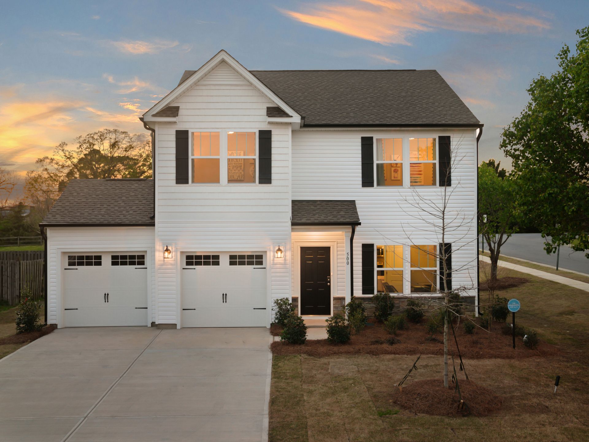 White two-story house with black shutters, a dark door, and a two-car garage on a lawn at sunset.