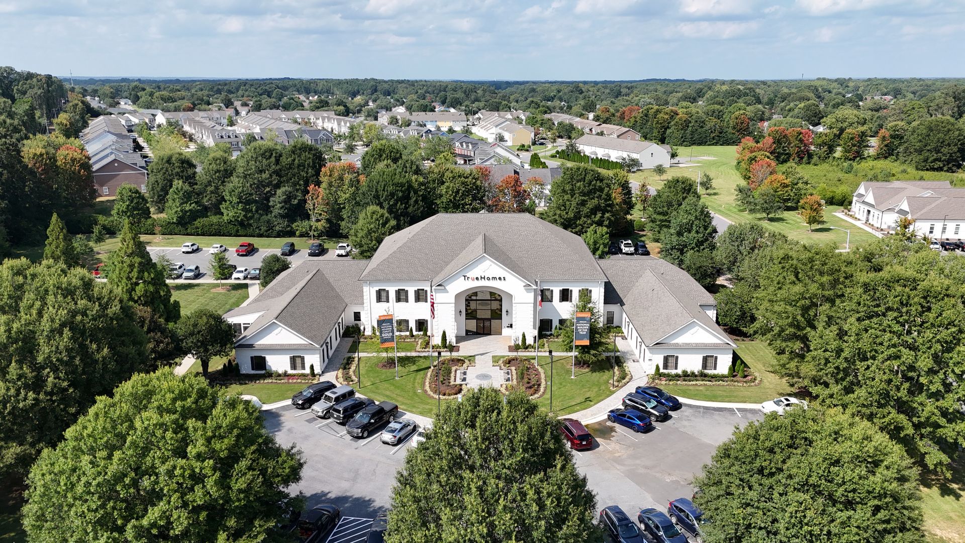 Aerial view of a white, multi-winged building surrounded by green trees and a parking lot; residential area