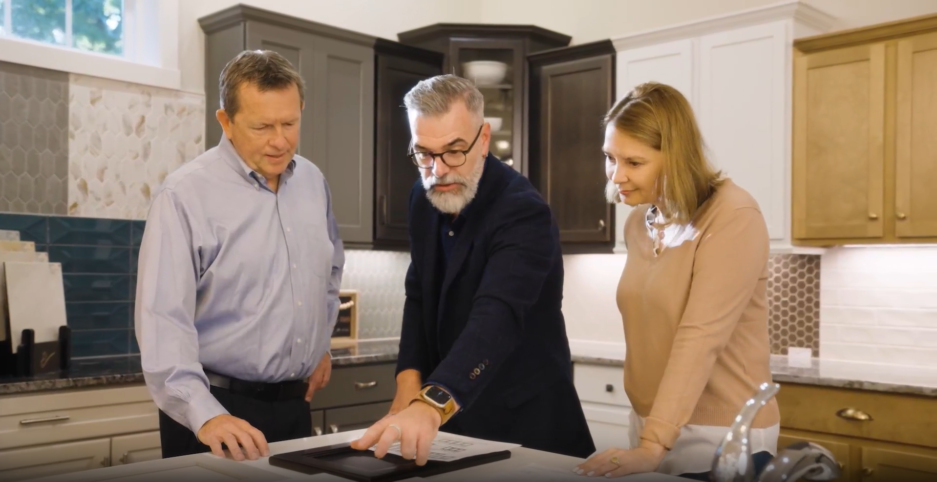 Three people in a kitchen discussing design options. A man points to a countertop, two others look on, cabinets