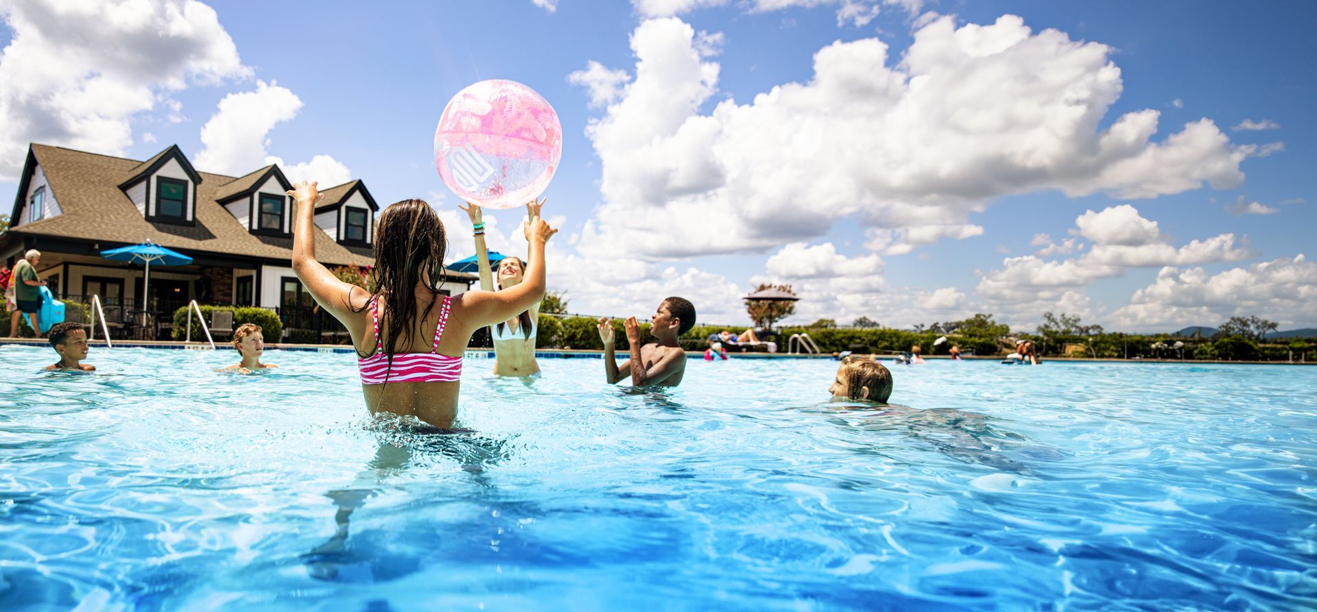 Children play in a large pool on a sunny day. One girl holds a pink beach ball. Blue water, sky, and building.