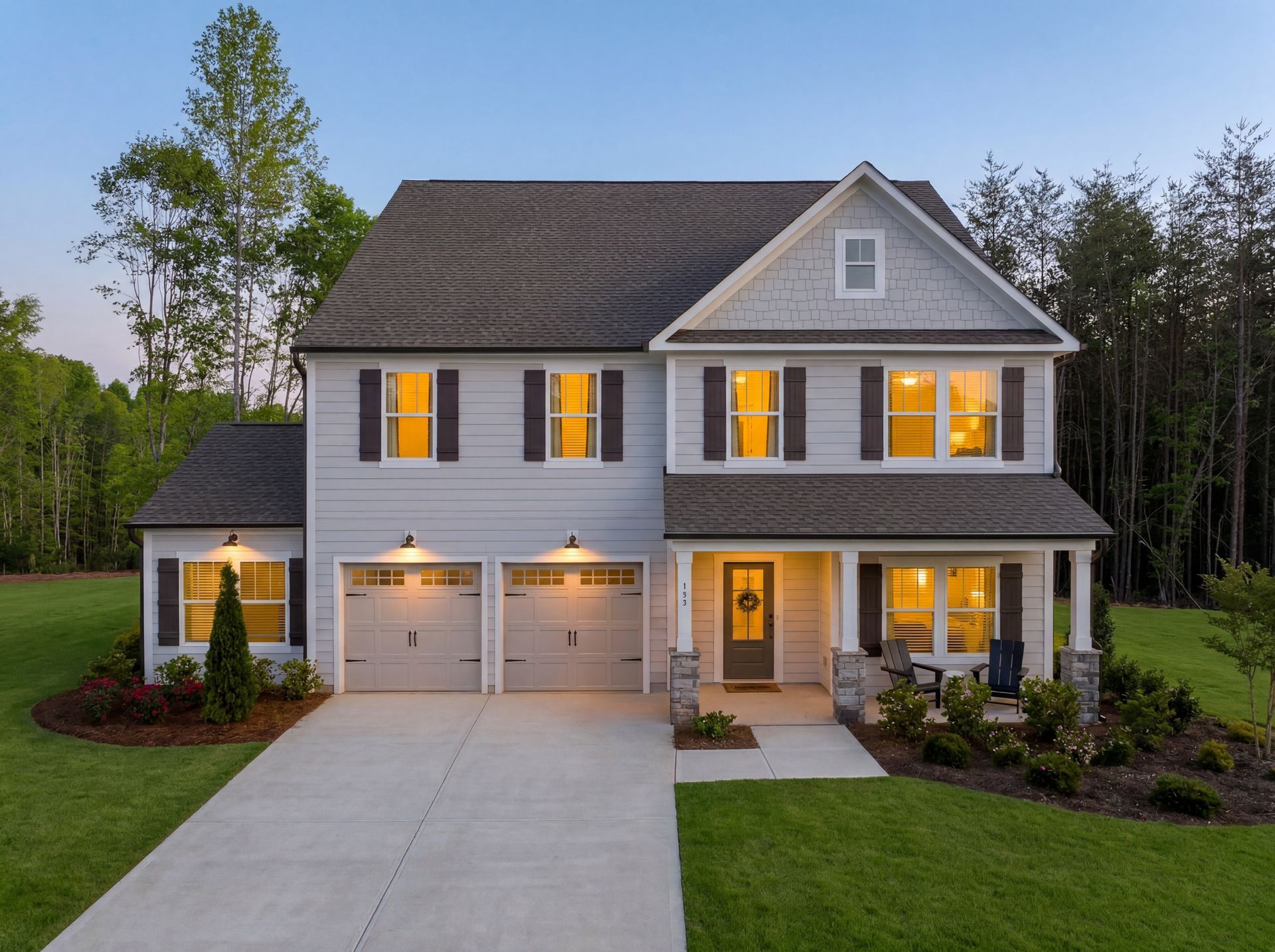Two-story gray house with lit windows, double garage, front porch, and landscaped lawn at dusk