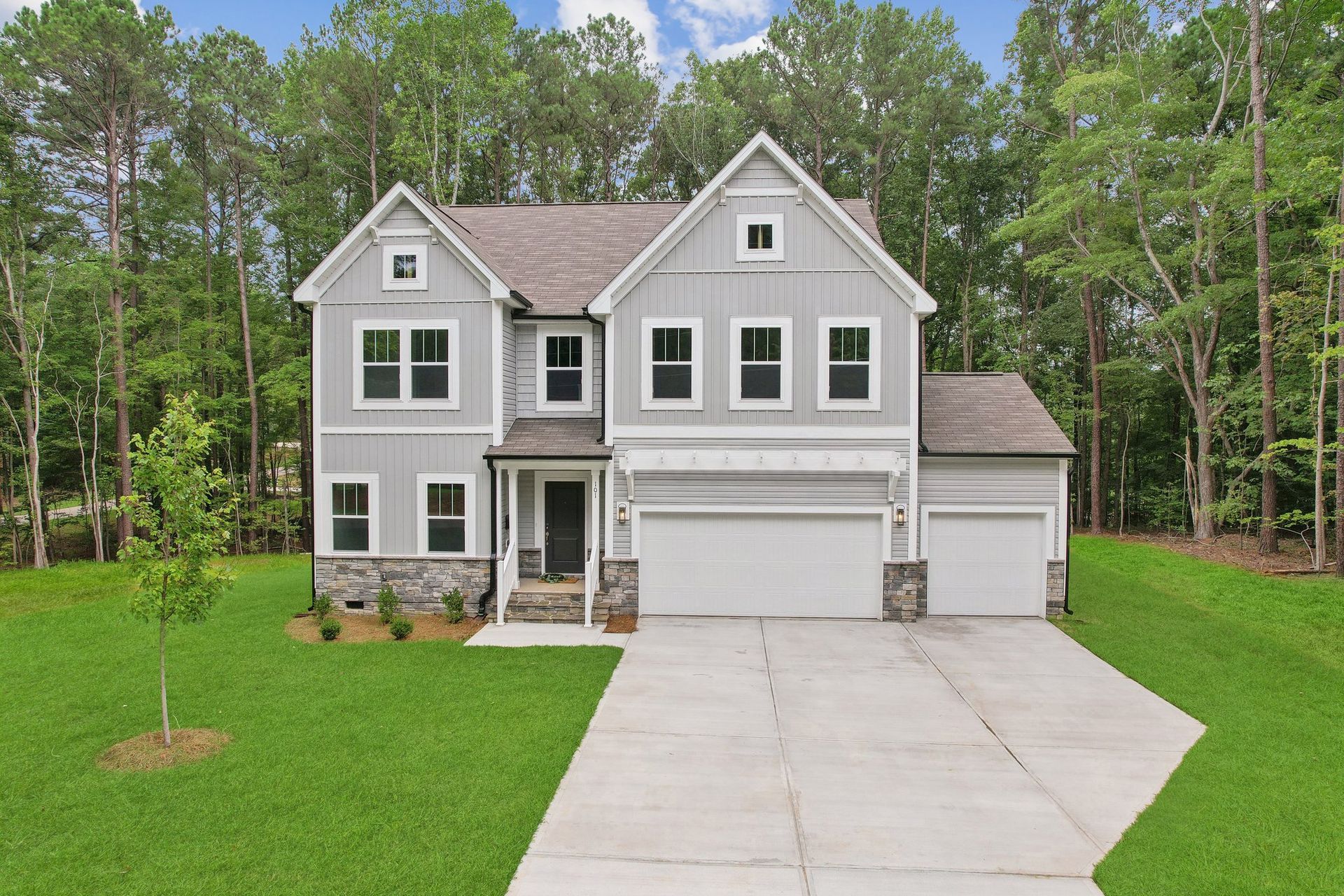 Two-story gray house with white trim, stone accents, and attached garage, set against a backdrop of trees.