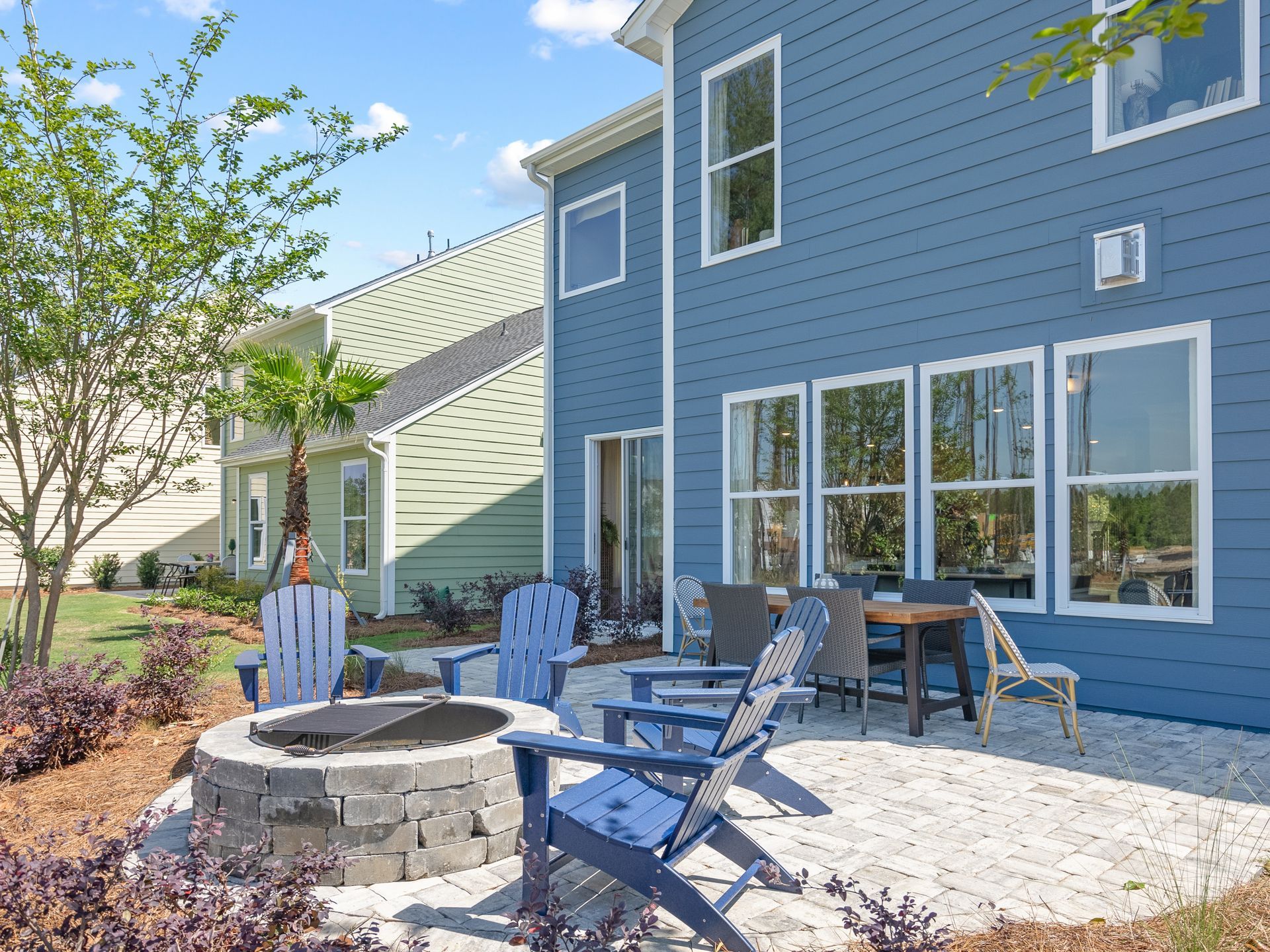 Backyard patio with blue Adirondack chairs, fire pit, and a table next to a teal house.