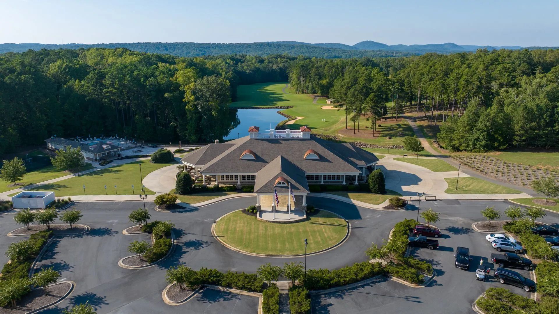 Golf course clubhouse surrounded by trees and a pond, with a circular driveway and parking area.