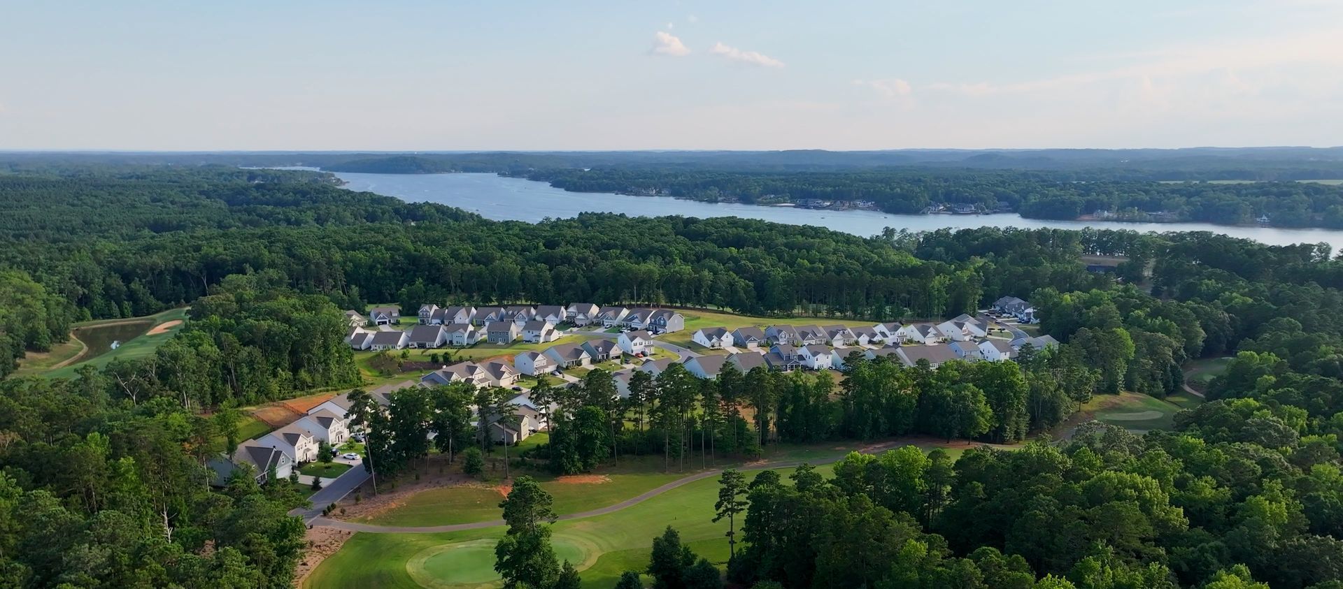 Aerial view of a residential area surrounded by lush green trees, with a lake in the distance.