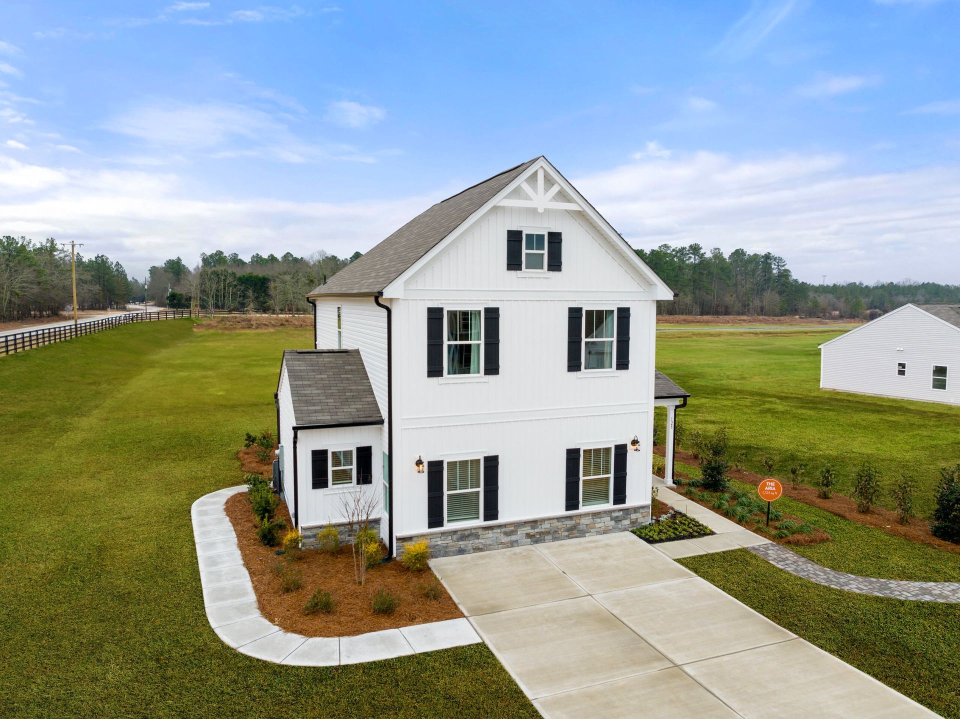 White two-story house with black shutters, driveway, and small building in a green field on a sunny day.